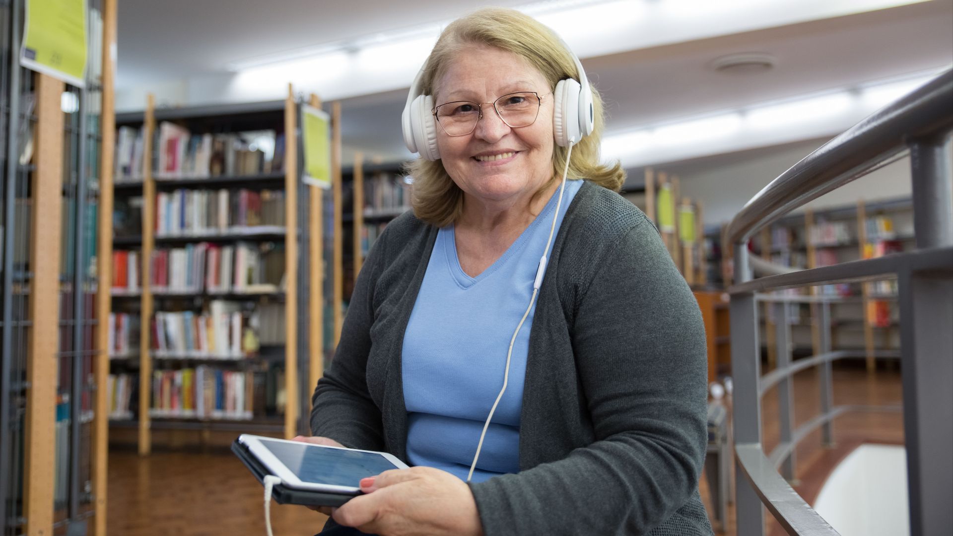 Senior woman smiling while listening to audiobooks on tablet in library setting.