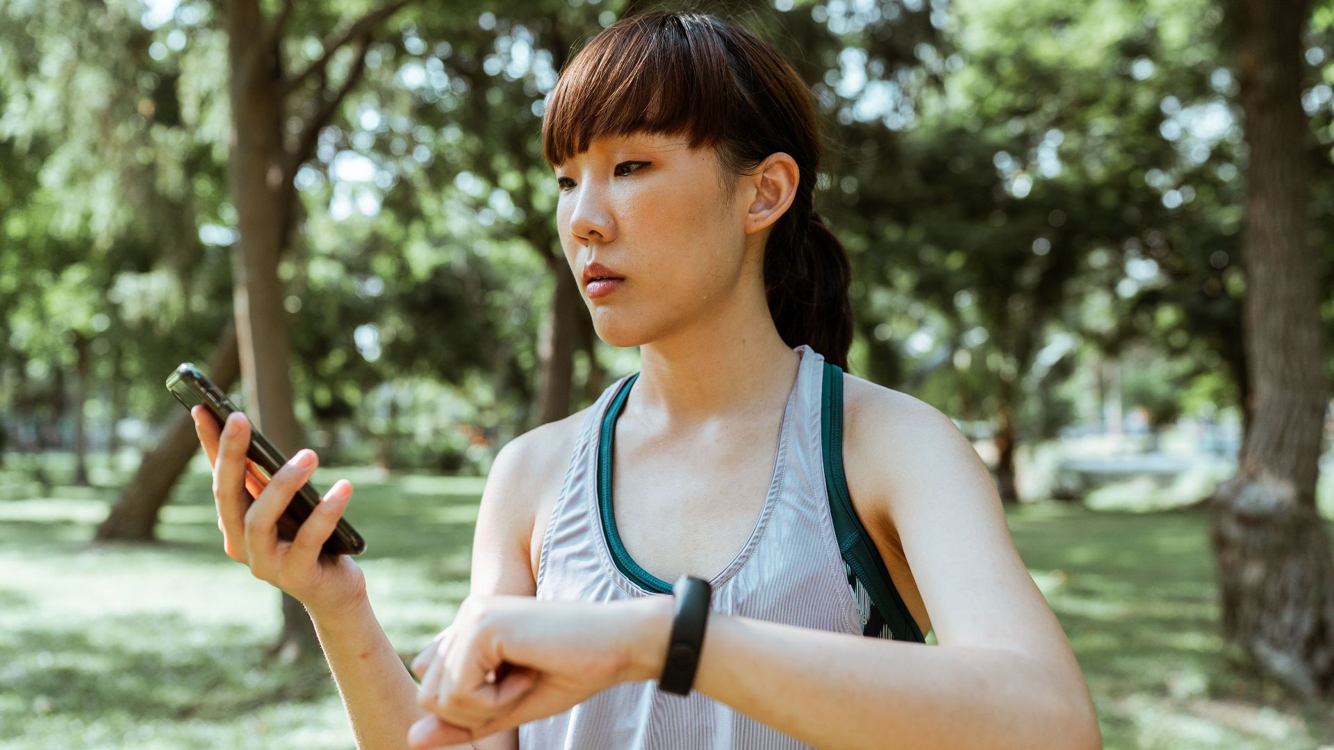 Serious young Asian female in sportswear using smartphone and smart watch in park on sunny summer day