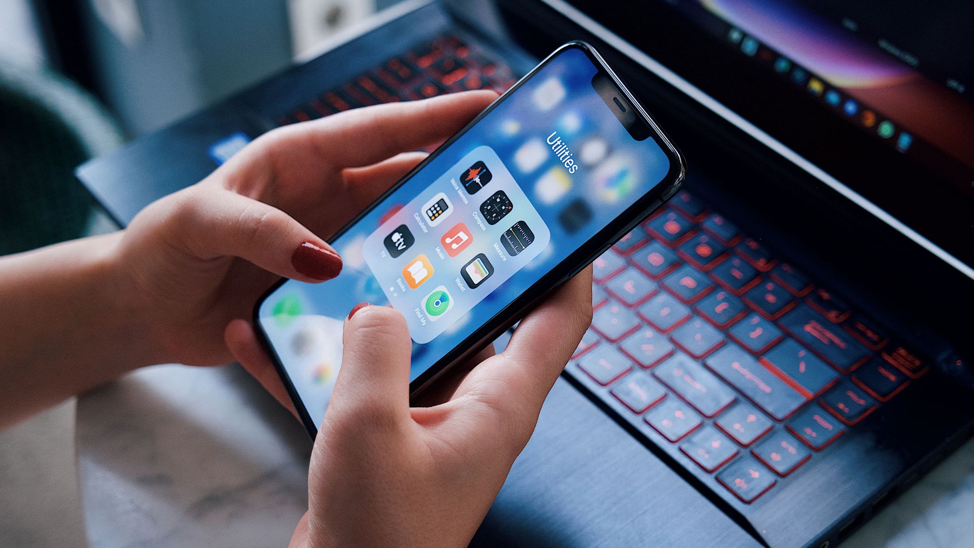 Close-up of a woman holding a smartphone showing utilities apps while sitting by a laptop indoors.