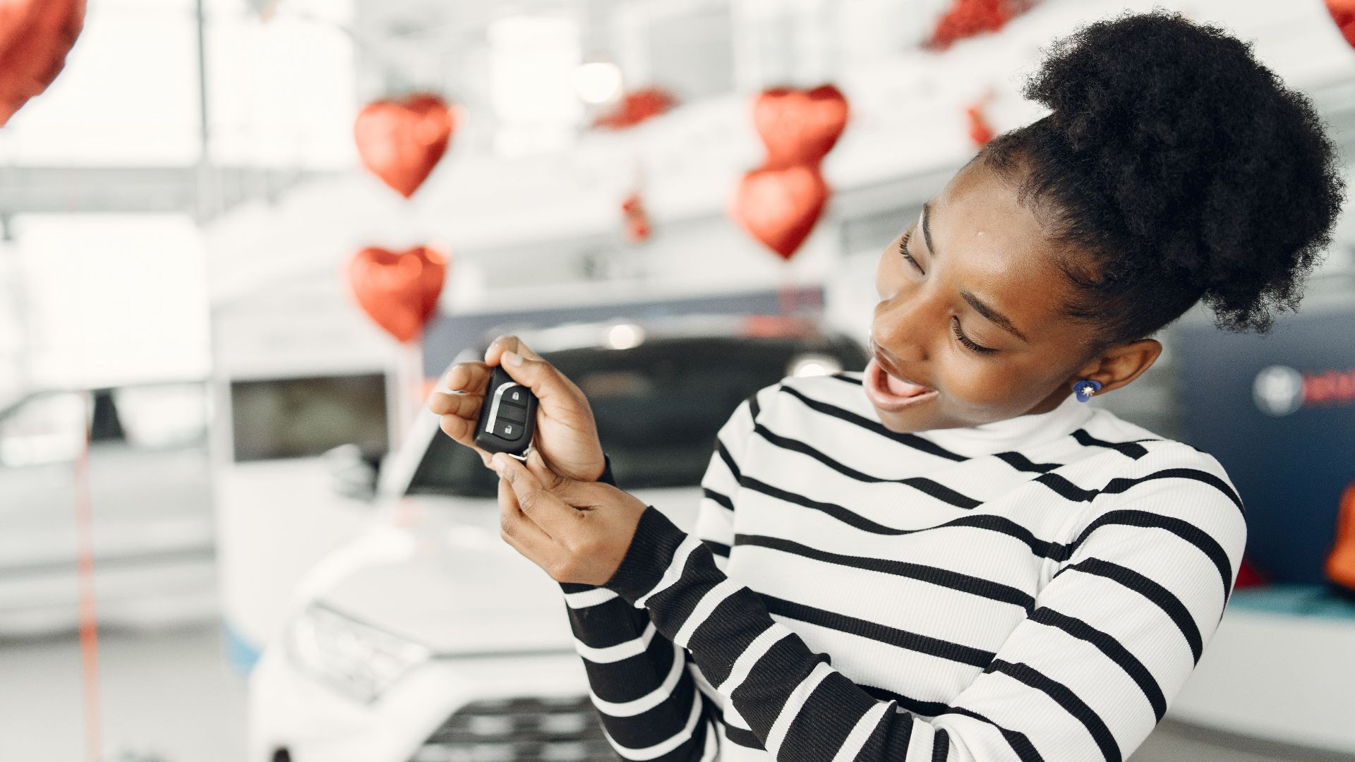 Joyful woman holding car key amidst heart decorations in a showroom.