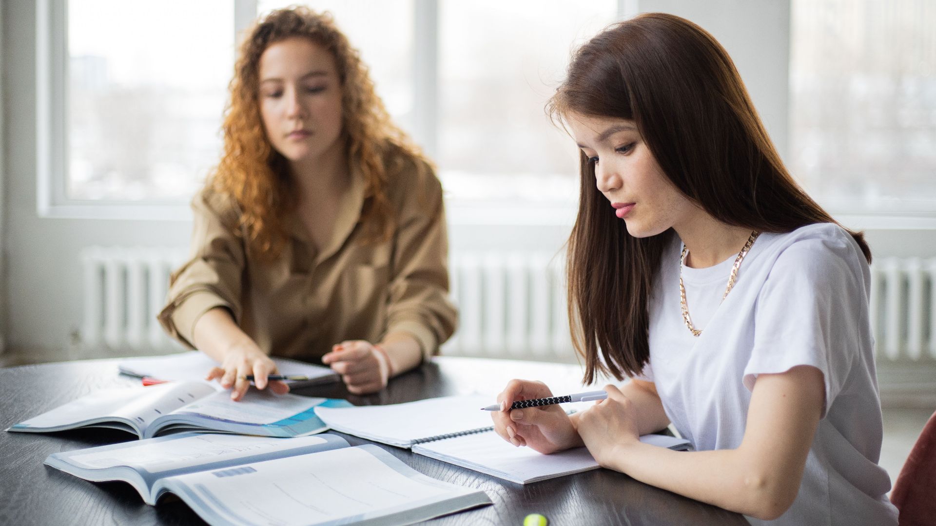 Serious multiracial female students writing in notebooks at table with textbook while preparing for exam together in classroom during lesson