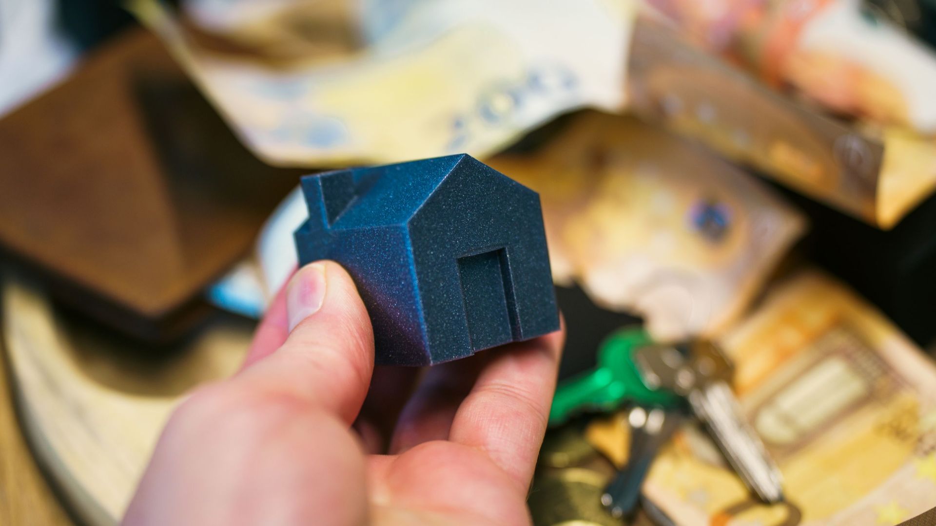 Close-up of a hand holding a small house model surrounded by euro banknotes and keys, symbolizing real estate investment.