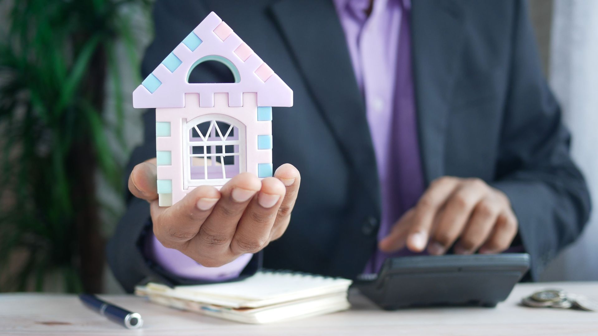 Businessman in suit holds model house, calculates real estate investments.