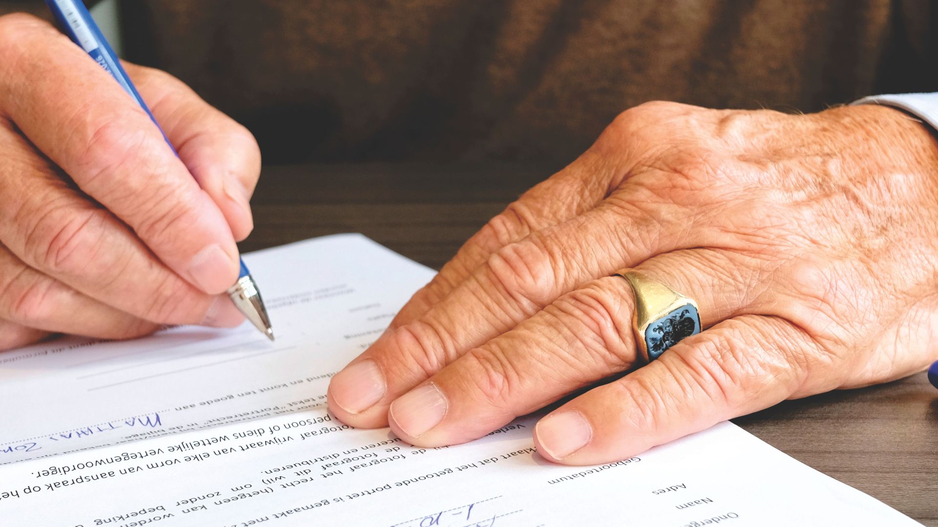 Close-up of a senior adult signing a legal document with a focus on hand and gold ring.