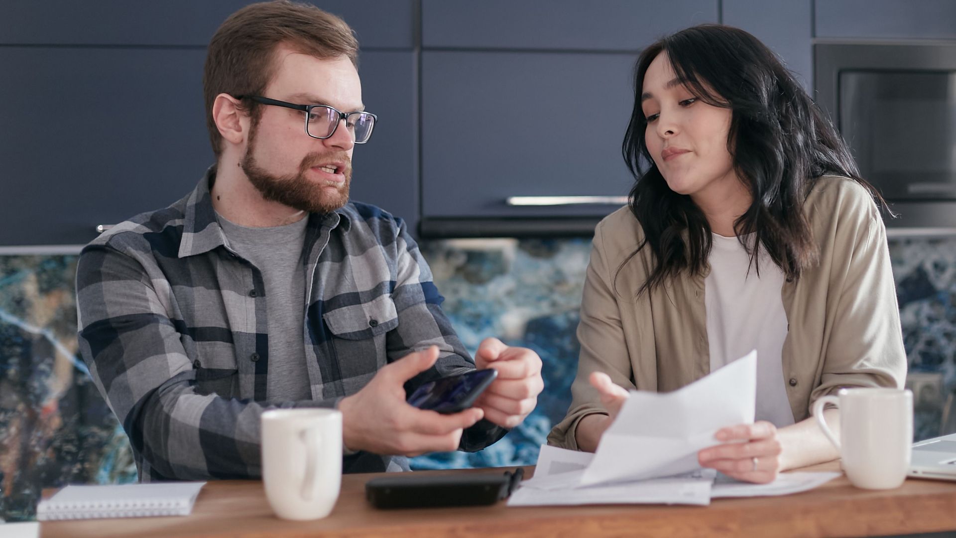 A couple sits at a kitchen table reviewing documents and discussing finances.