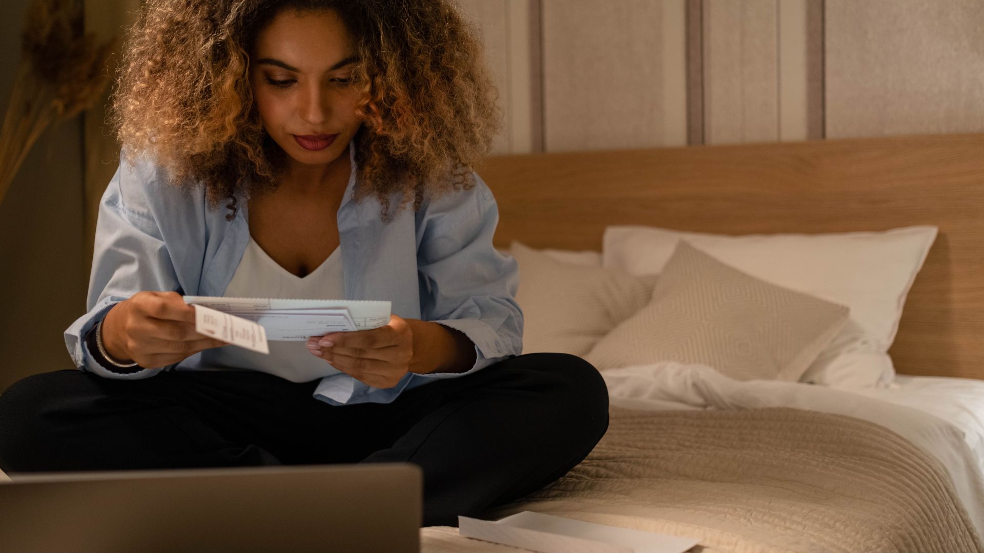 Woman sitting on bed reading documents with laptop, highlighting home office setting.
