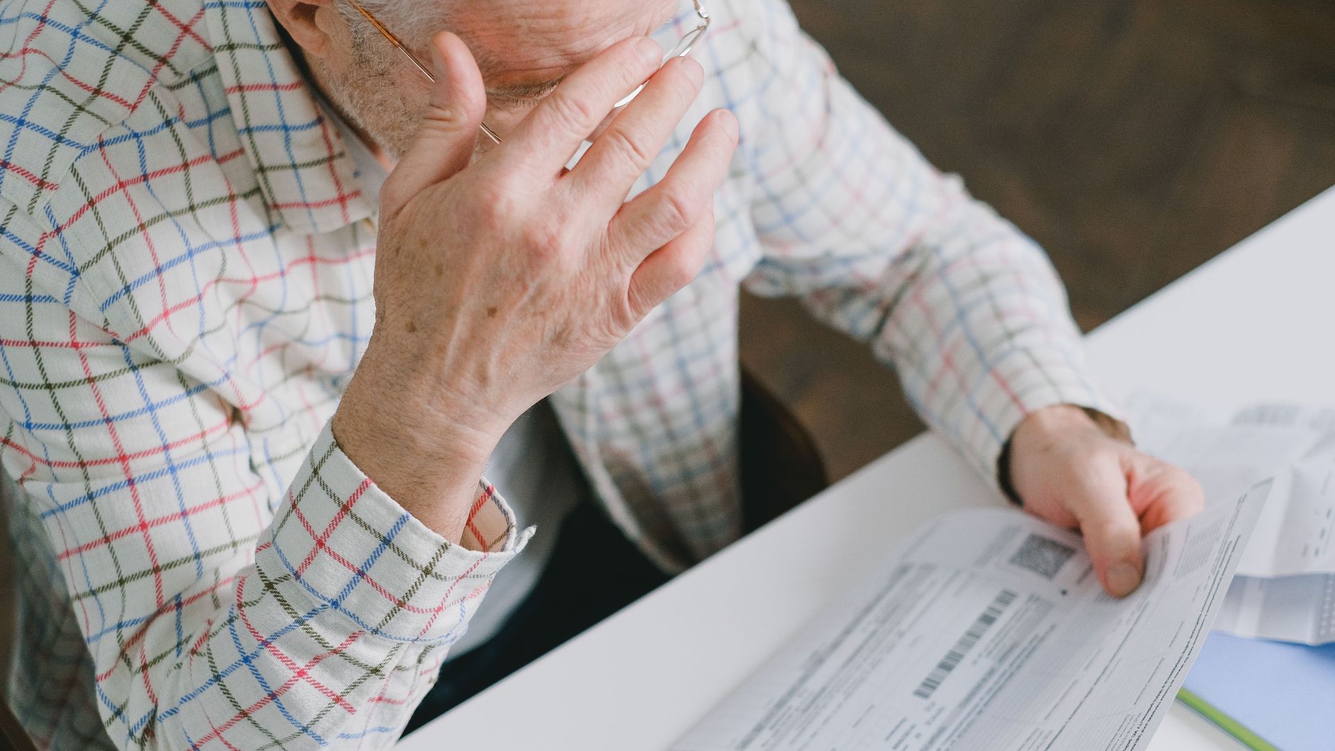 Elderly man in patterned shirt reading and holding bills at a home table, appearing focused.