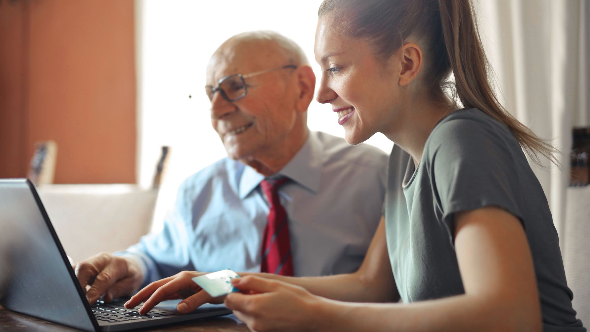 Young woman in casual clothes helping senior man in formal shirt with paying credit card in Internet using laptop while sitting at table