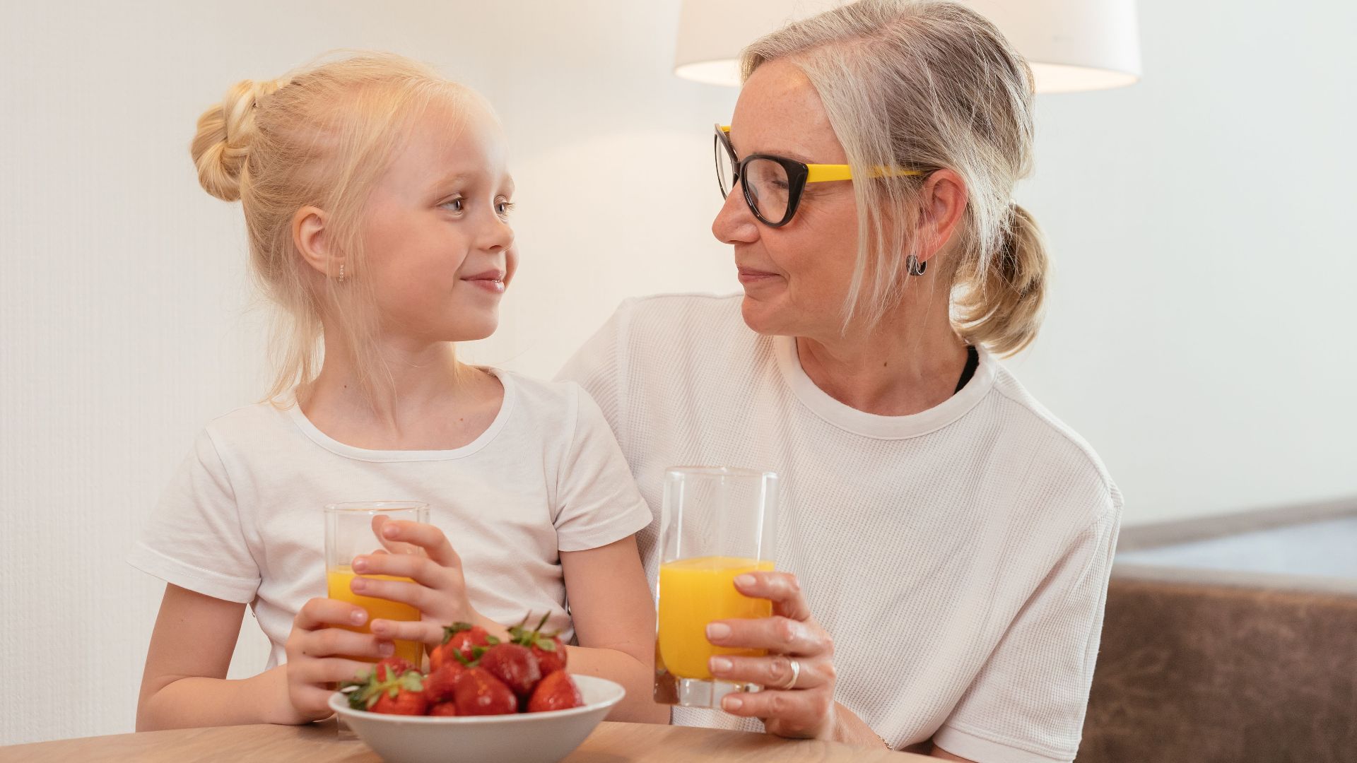 Elderly woman and young girl enjoying orange juice and strawberries at home, sharing a joyful moment together.