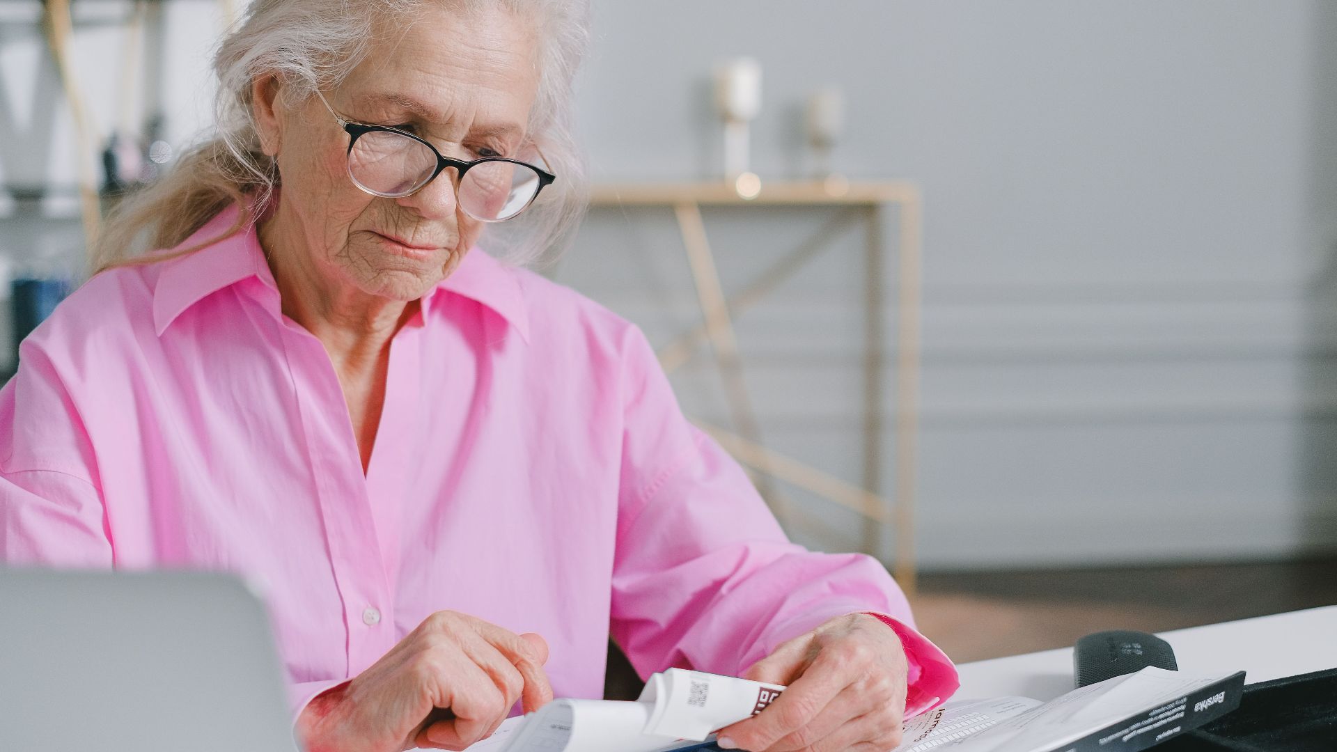 Elderly woman in pink shirt reading documents at a desk indoors.