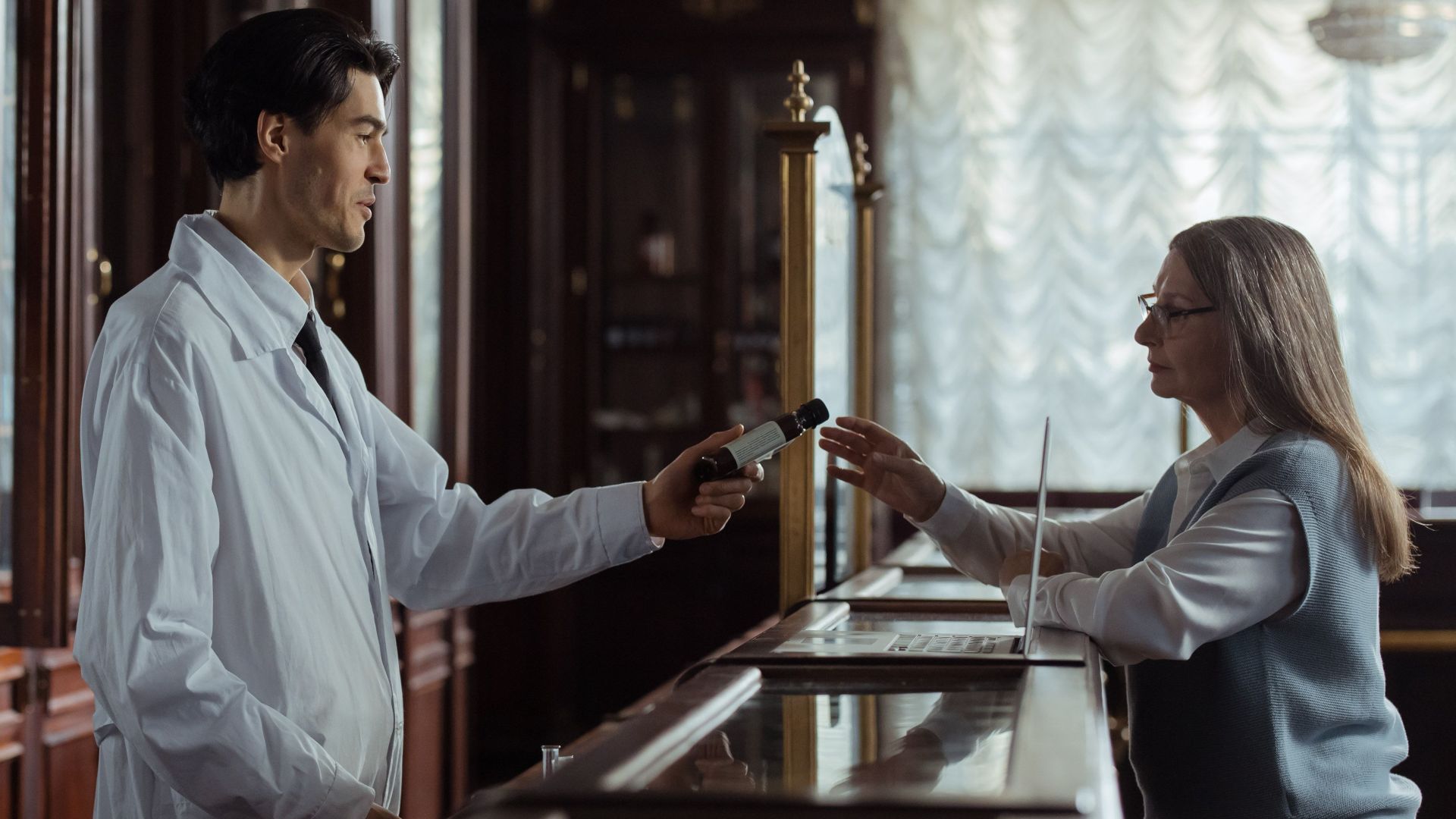A pharmacist hands medication to a customer at a classic vintage pharmacy counter.