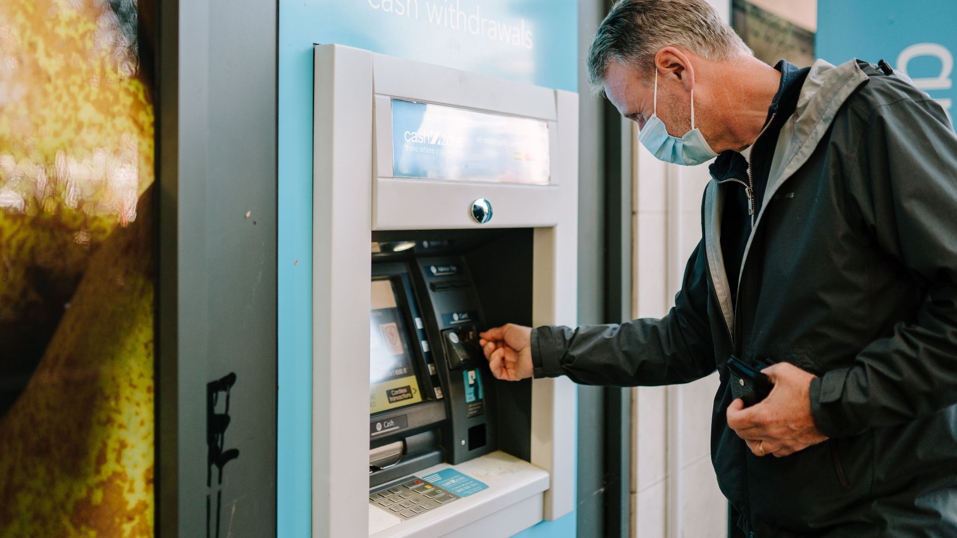 A man wearing a mask using an ATM machine outdoors for cash withdrawal.