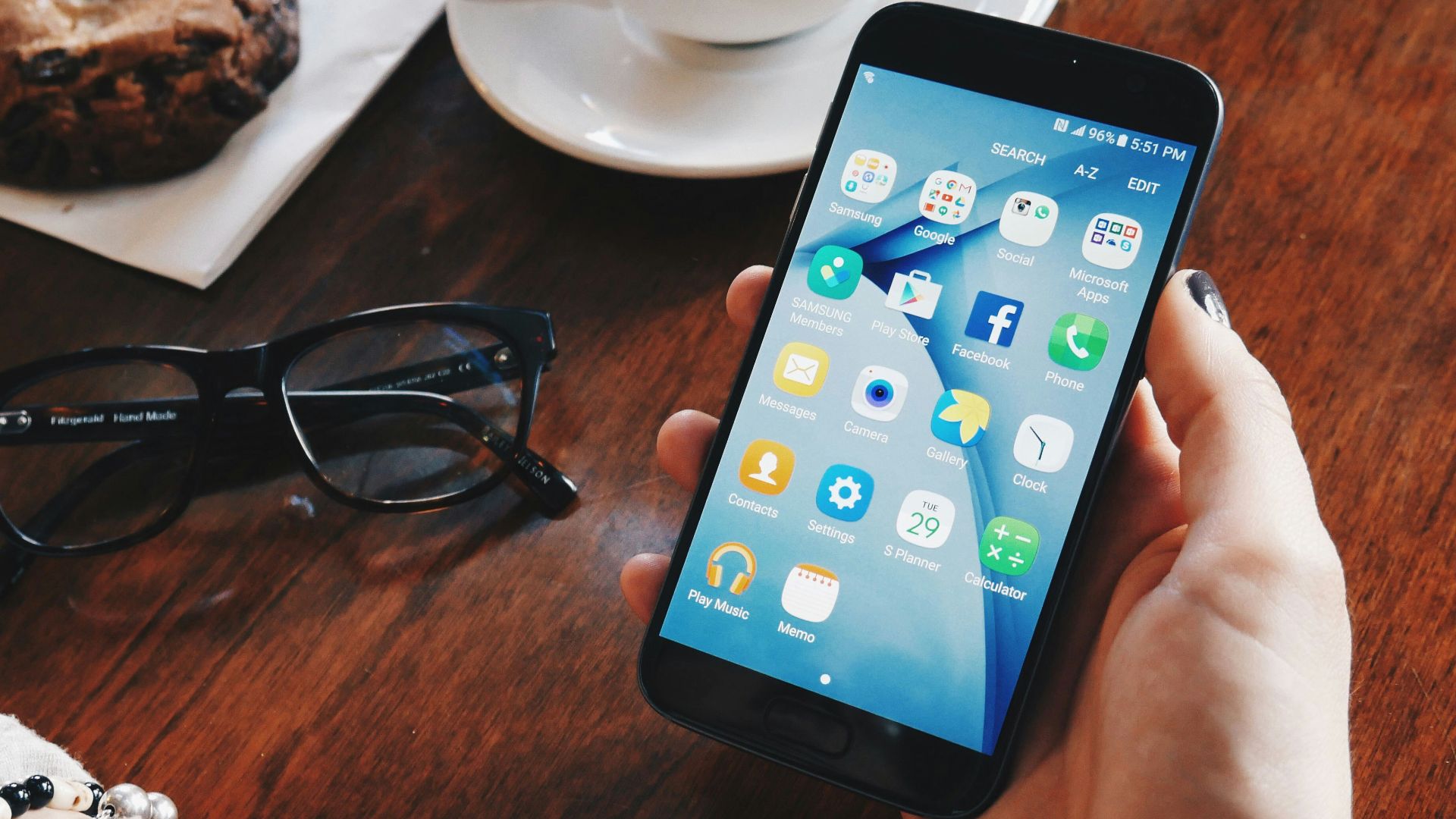 person holding turned-on smartphone sitting at table with filled cup with saucer