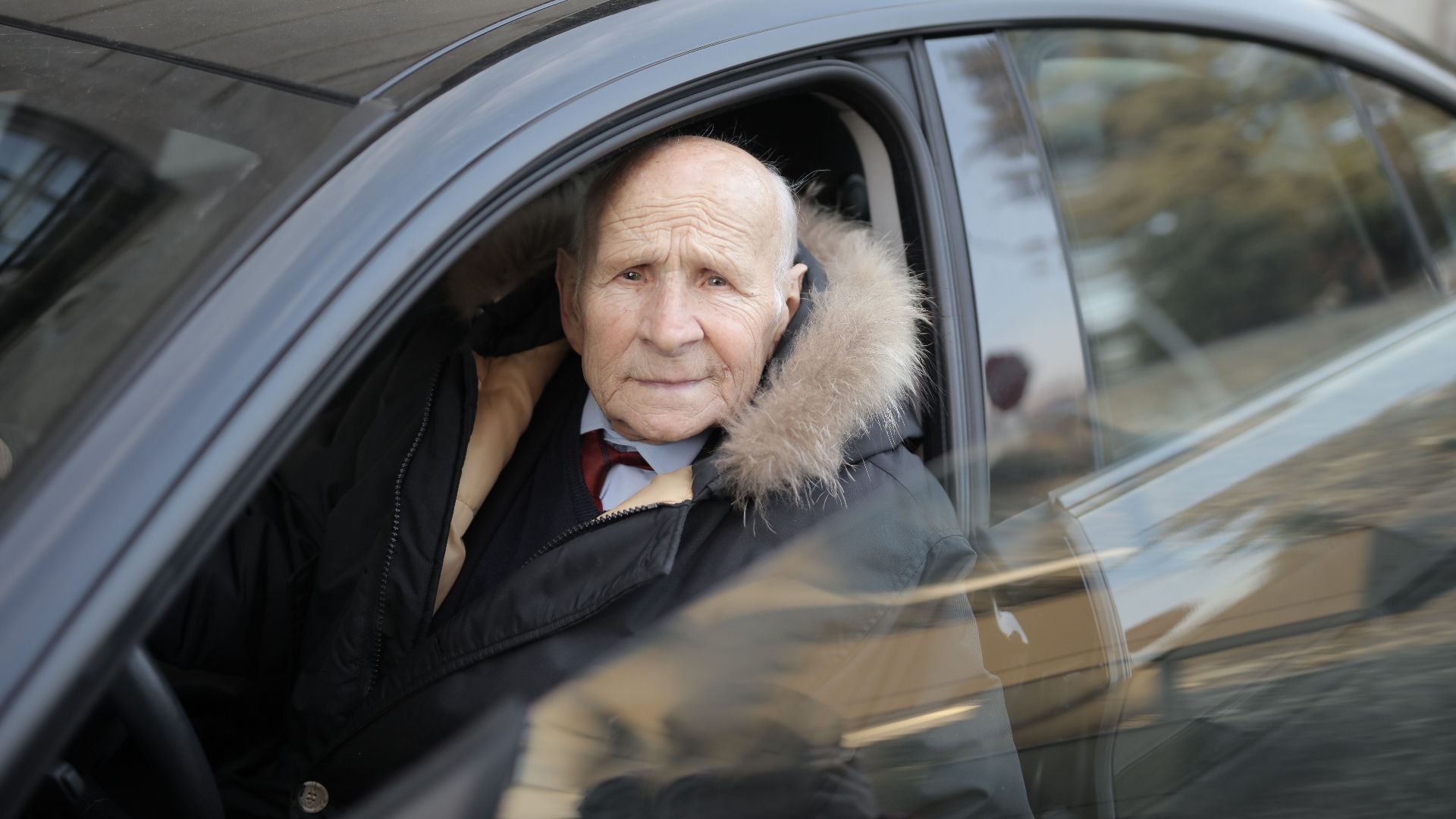 Senior man driving a car, wearing a winter coat, captured through the vehicle window.