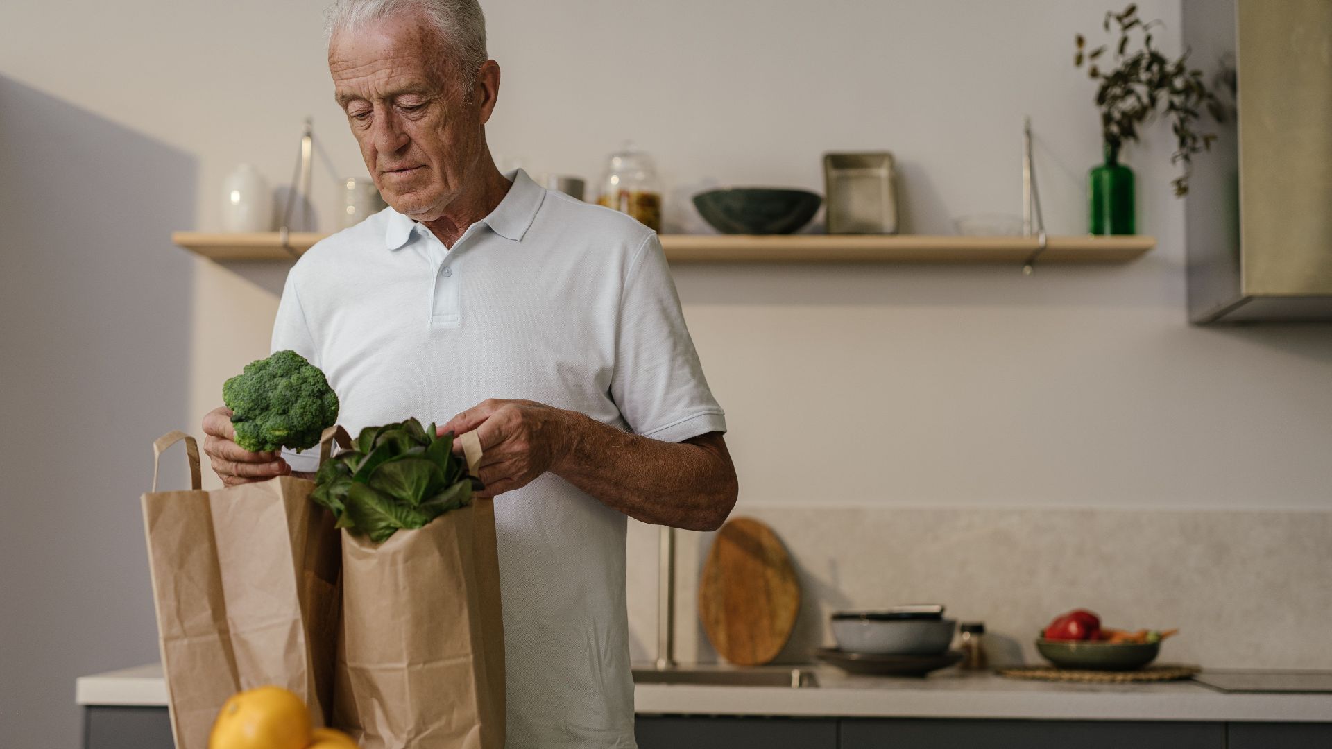 Elderly man in kitchen examining fresh vegetables from paper bags. Healthy lifestyle concept.