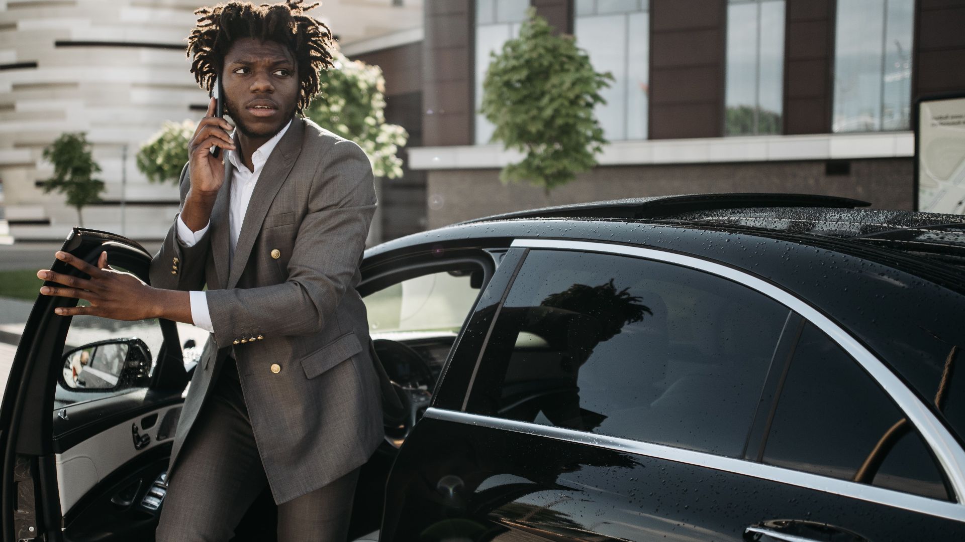 Confident businessman in a suit talking on a smartphone while getting out of a luxury black vehicle.