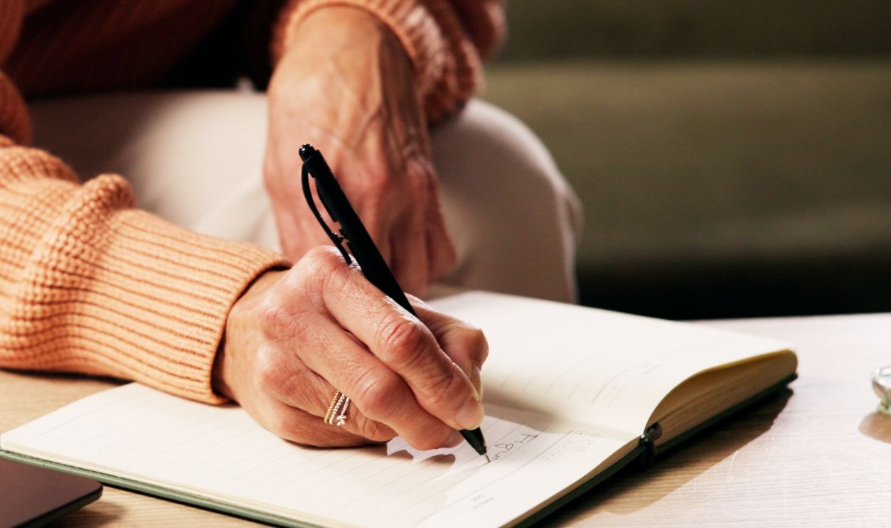 Hands, person and writing on journal in home for reminder, agenda or planning with ideas. Diary, notes and senior woman with notebook for checklist, tasks and schedule with information for to do list