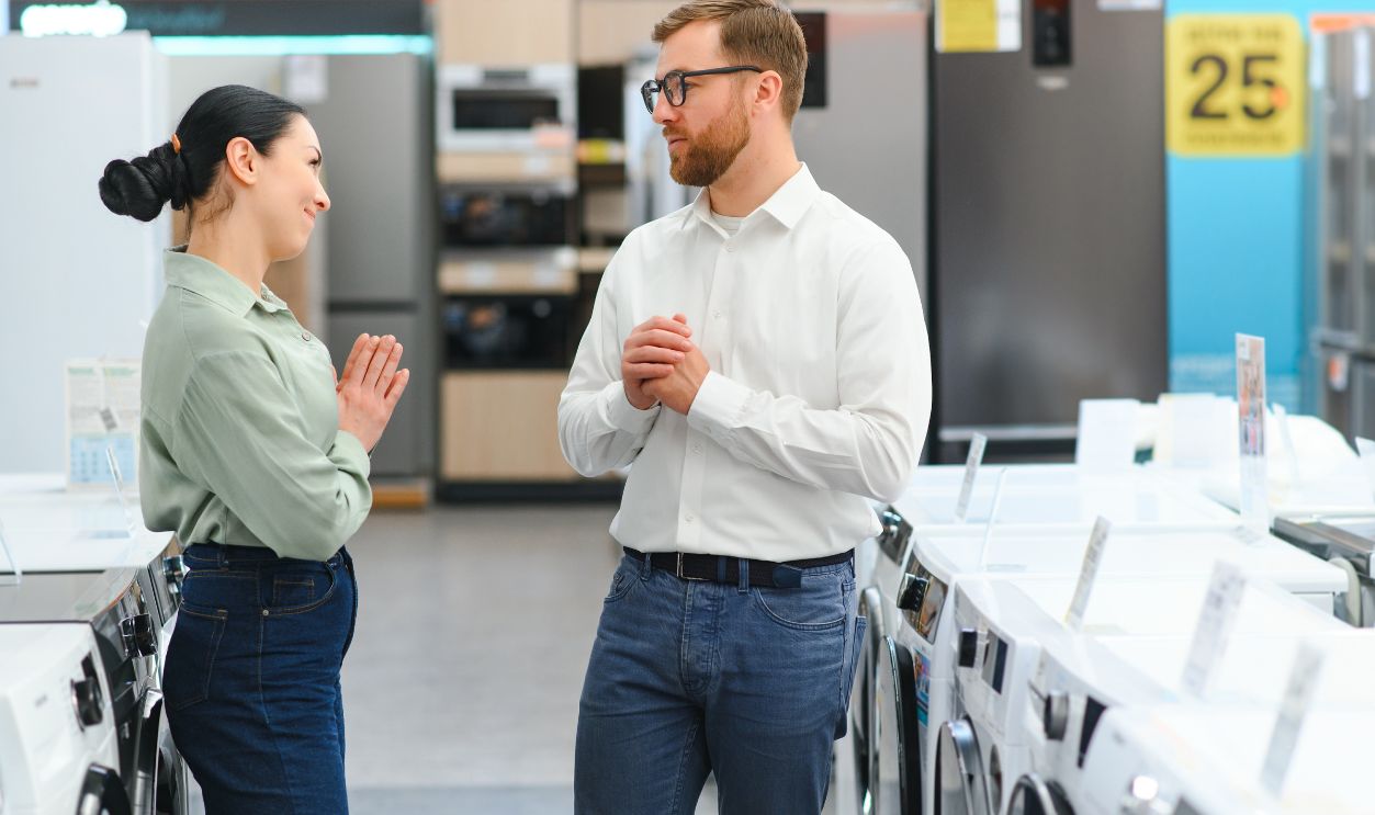 Portrait of a Retail Electronics Shop Consultant Assisting Young Female Customer in Selecting a Washing Machine For Her Home. Beautiful Female Evaluating Modern Laundry Solutions