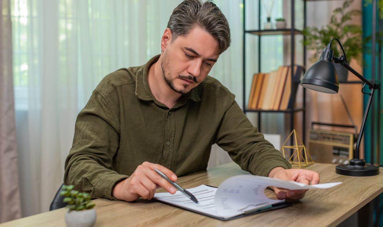 Shutterstock-2666798723, Middle-aged man at home reads contract document thinking deeply weighing decision feeling cautious. Young adult guy at table taps pen hesitates over legal terms before signing important agreement now