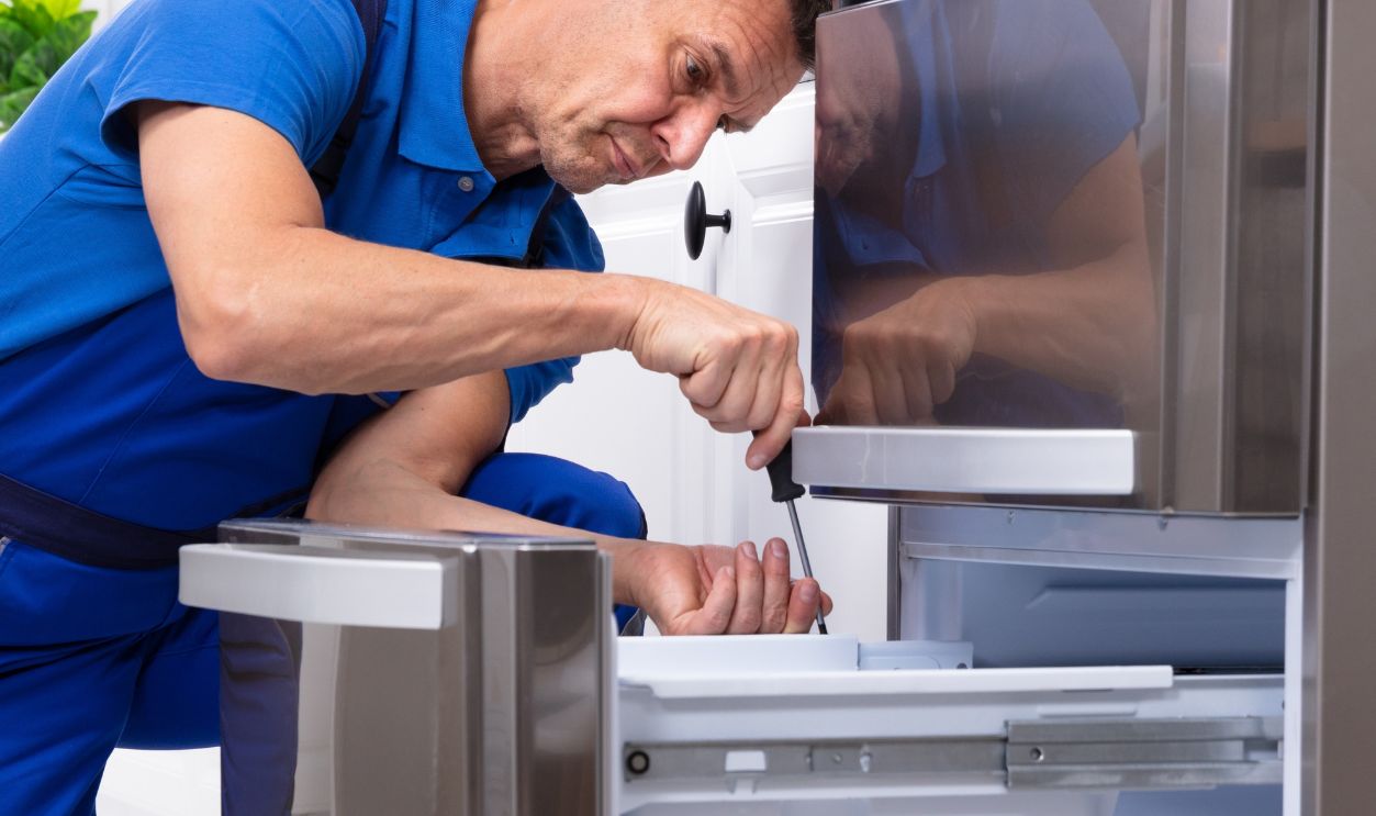 Mature Male Serviceman Repairing Refrigerator With Toolbox In  Kitchen