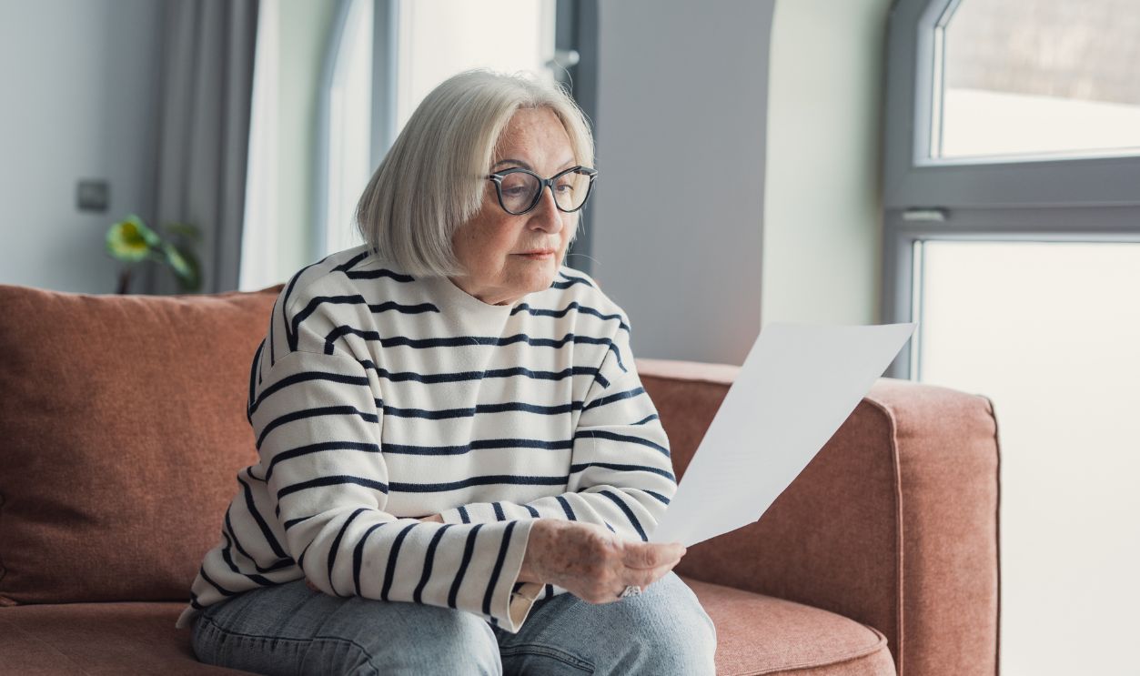 Focused elderly 60s woman sit on sofa in living room reading postal correspondence or bank notice, concentrated mature 50s female rest on couch at home consider message in paper post letter