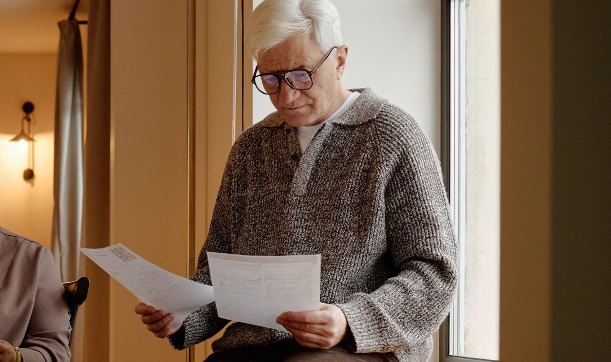 Senior Black woman and senior Caucasian man reading documents in modern kitchen, both sitting and focusing on paperwork, managing service bills together in natural light coming through large window