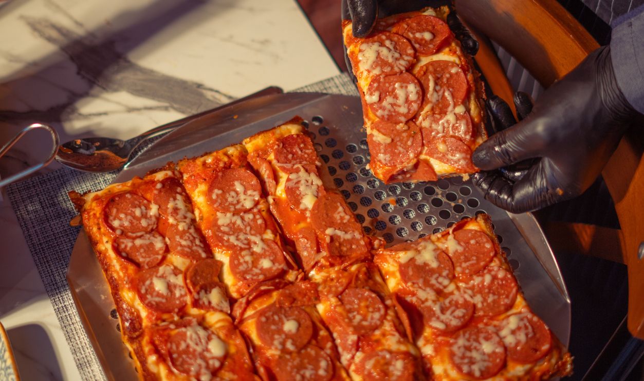 Chef serving square pepperoni pizza slices on metal tray in restaurant setting.