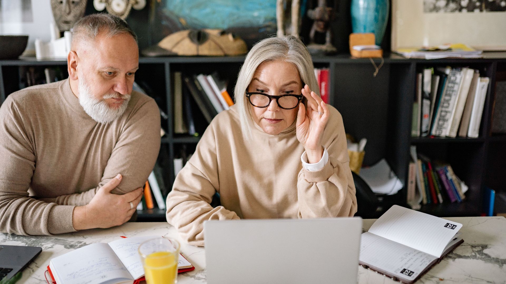 An elderly couple in a cozy home setting engaged with a laptop, planners, and a glass of juice on the table.