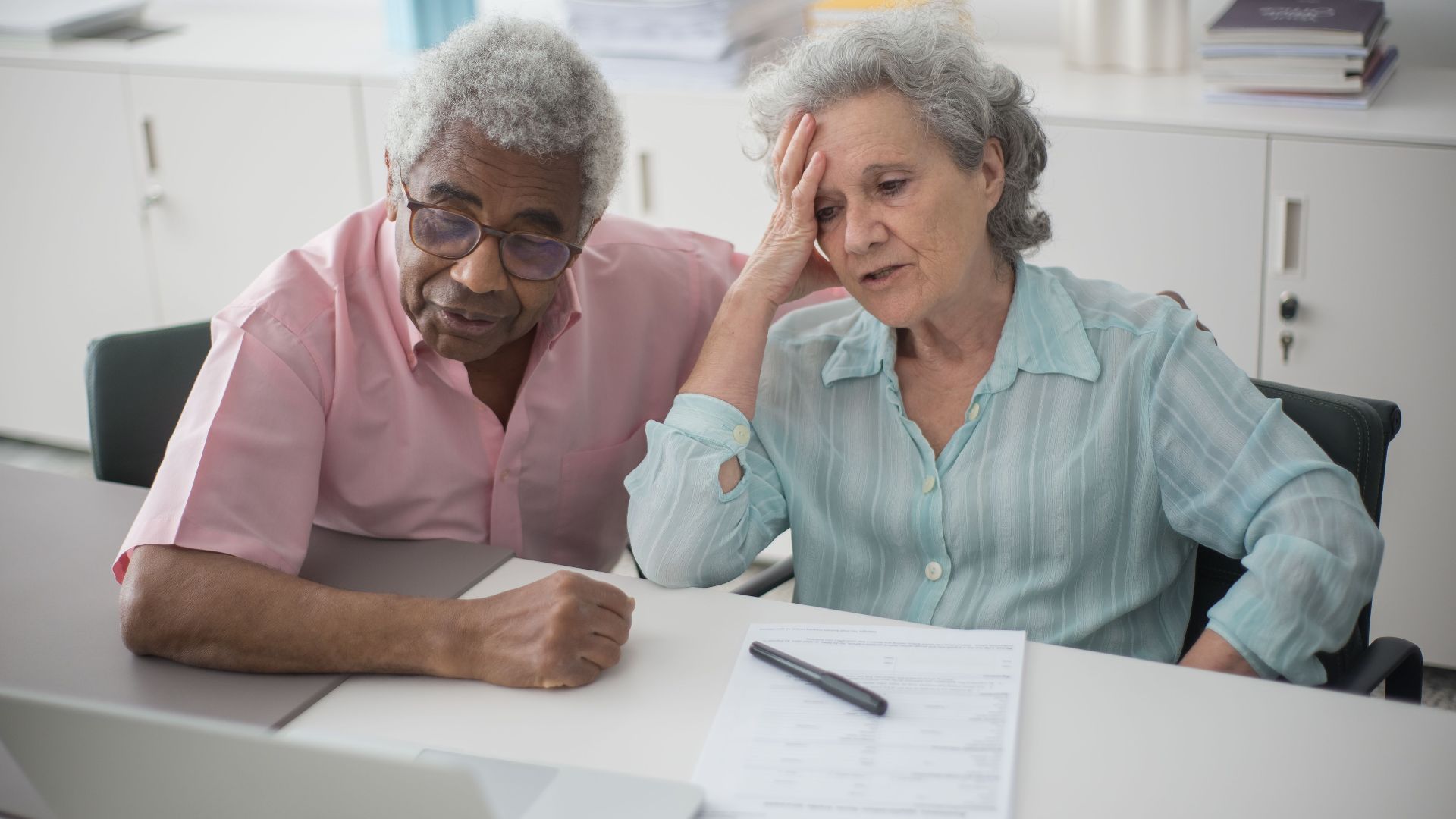 Elderly couple engaged with a laptop, reviewing paperwork at home office desk.