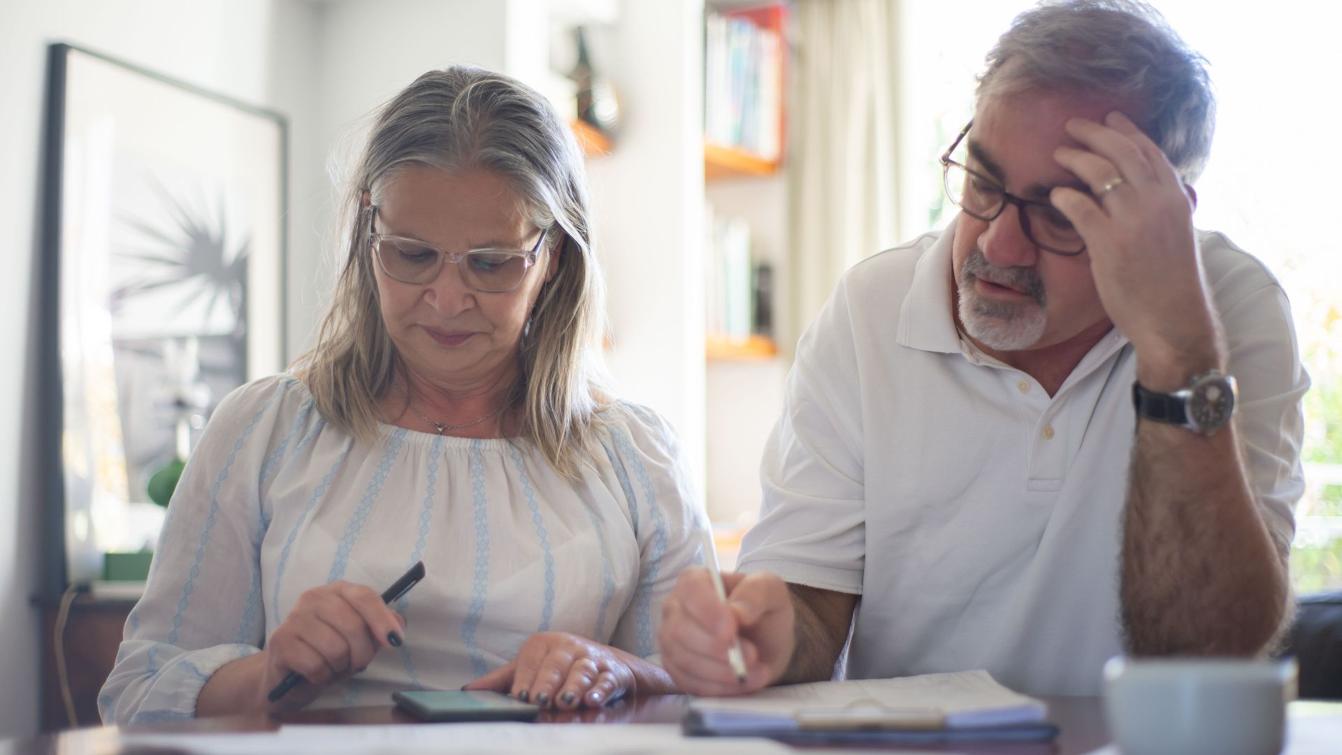 Senior couple calculating expenses at home office desk with documents and notes.