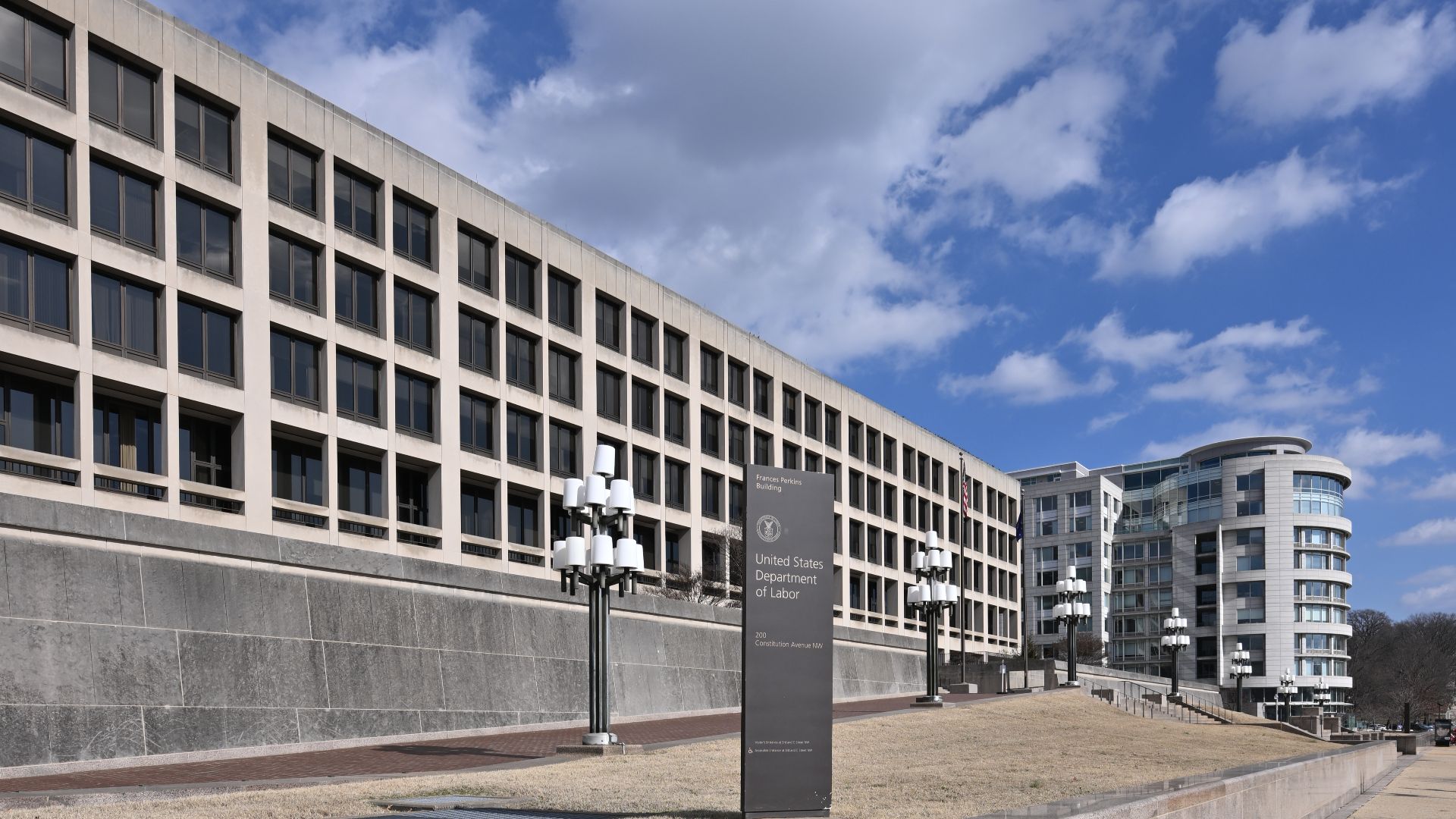 A sign on Constitution Ave for the United States Department of Labor with the Frances Perkins building behind it. 200 Constitution Avenue NW, Washington, DC 20004.