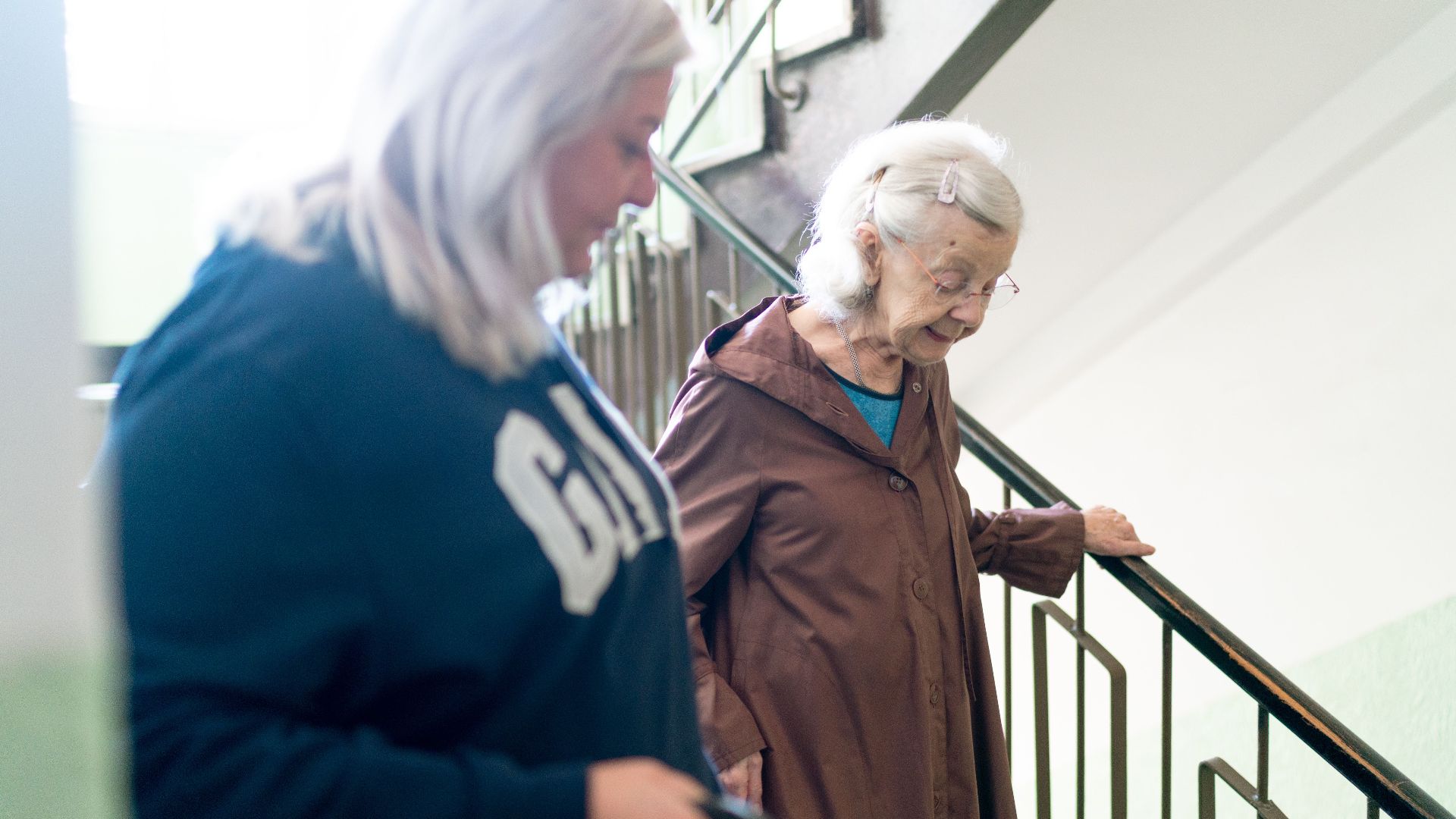 Elderly woman with caregiver on stairs, symbolizing home care and support, in Prague setting.