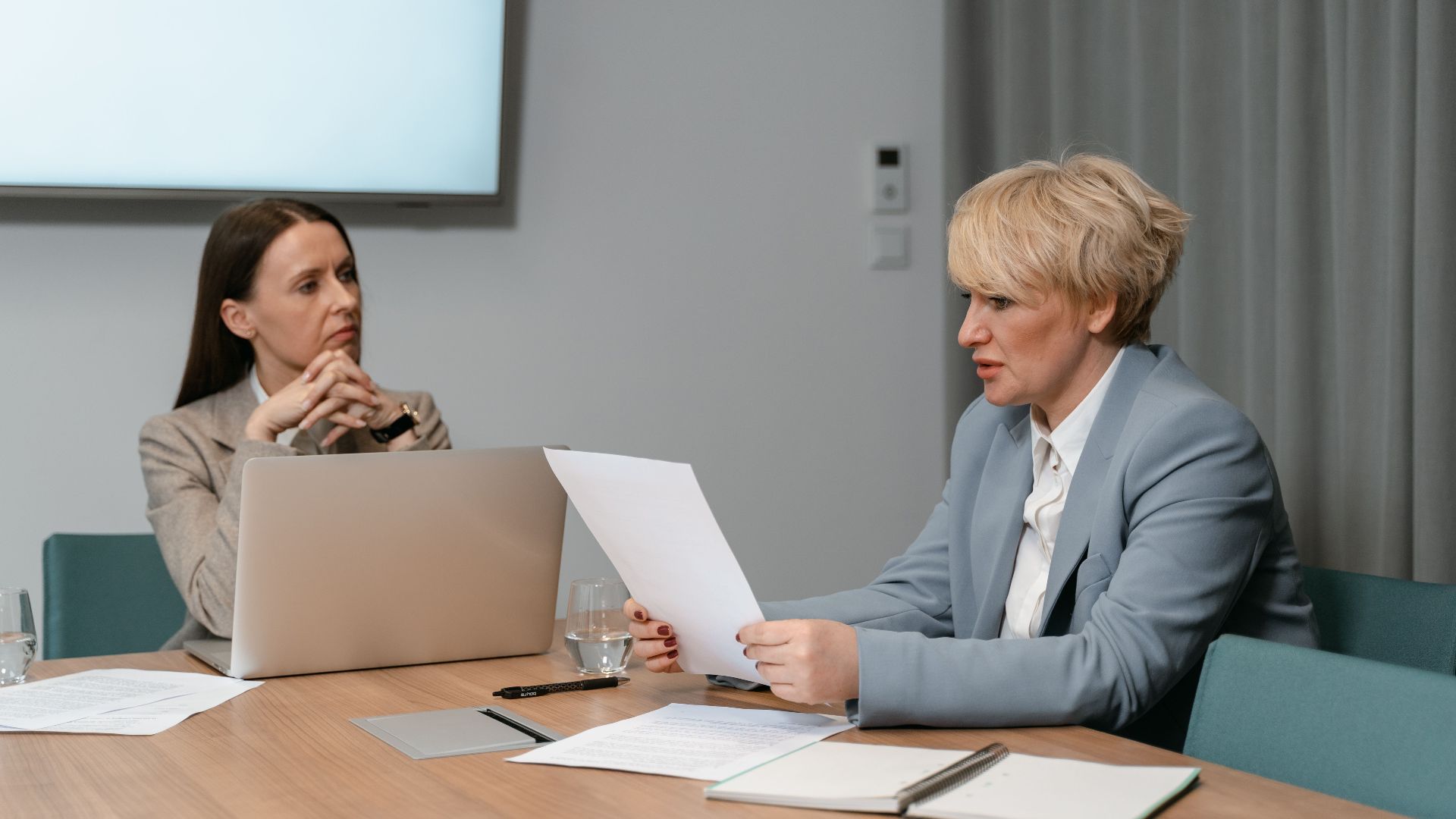 Two businesswomen in a serious meeting, discussing documents with a laptop in an office setting.