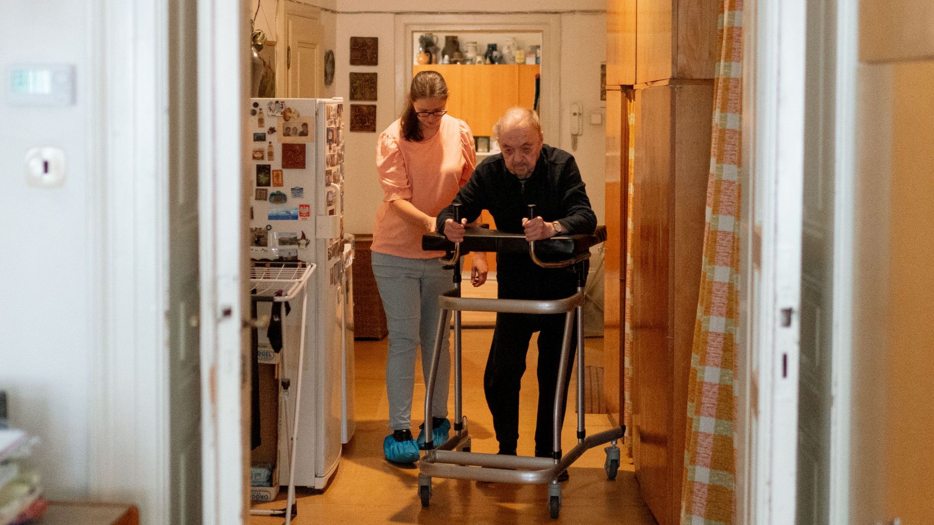 A caregiver assists an elderly man with a walker in a comfortable home environment.