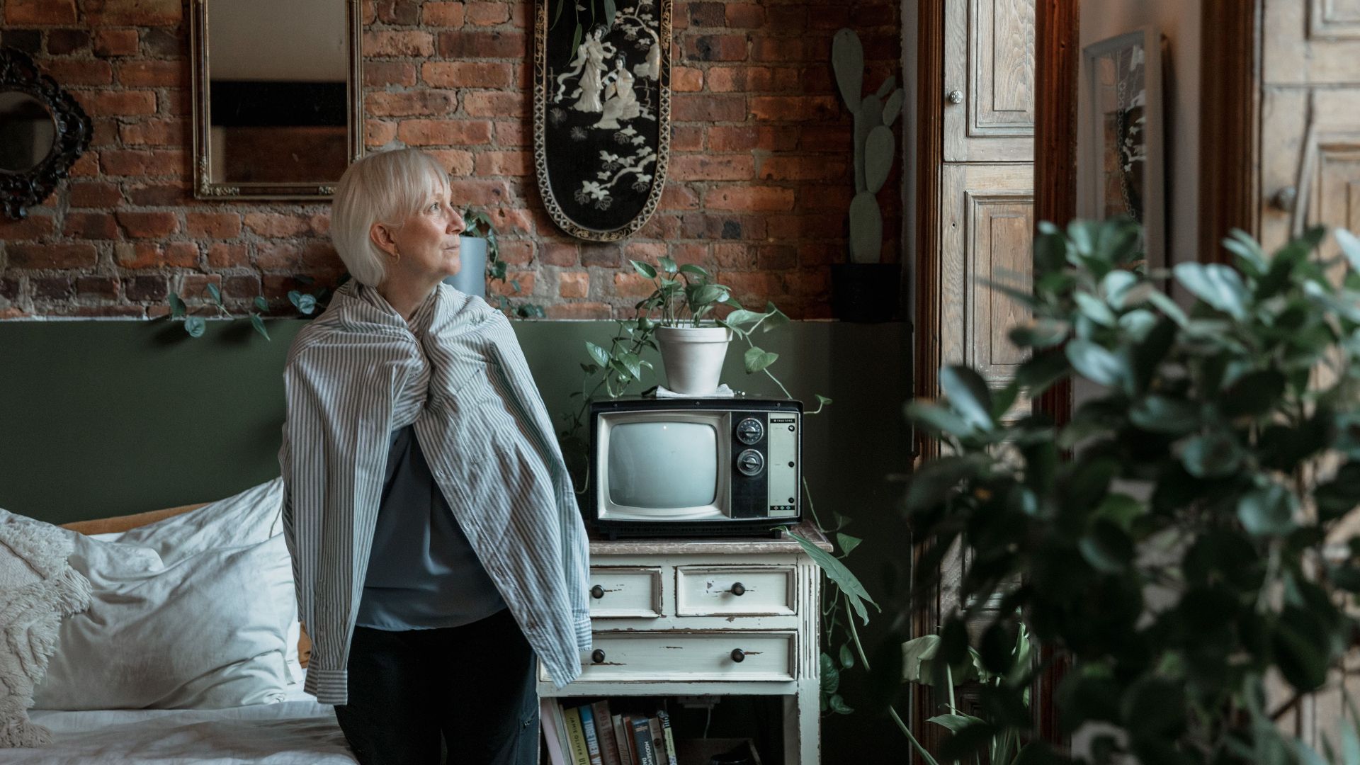 Elderly woman looks out window in cozy rustic room, surrounded by plants and vintage decor.