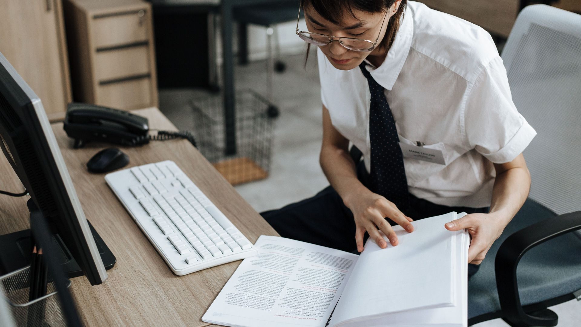 Professional in office attire reviewing documents at a desk with computer and book.
