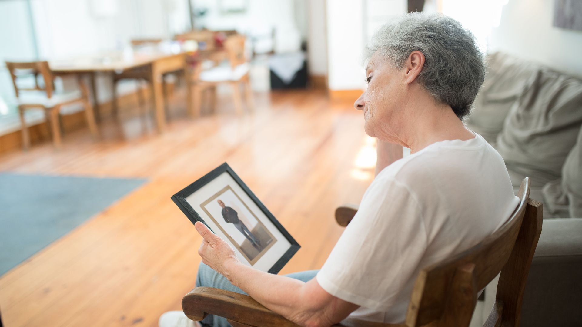 Senior woman gazing at photo frame in bright living room, conveying nostalgia and reflection.