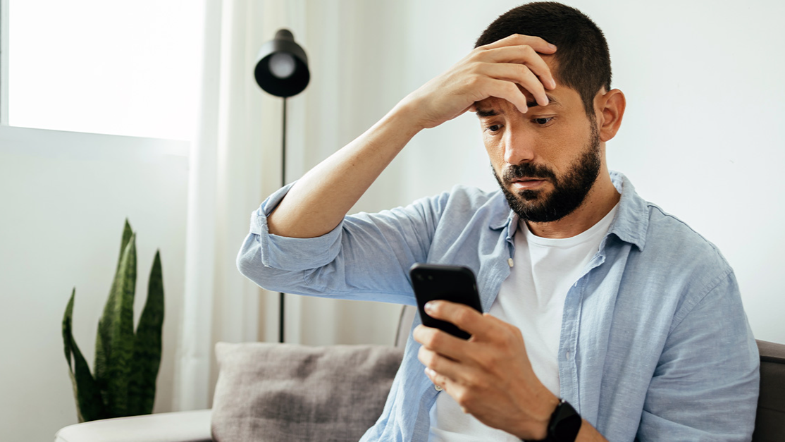 Sad man checking smartphone sitting on a sofa at home
