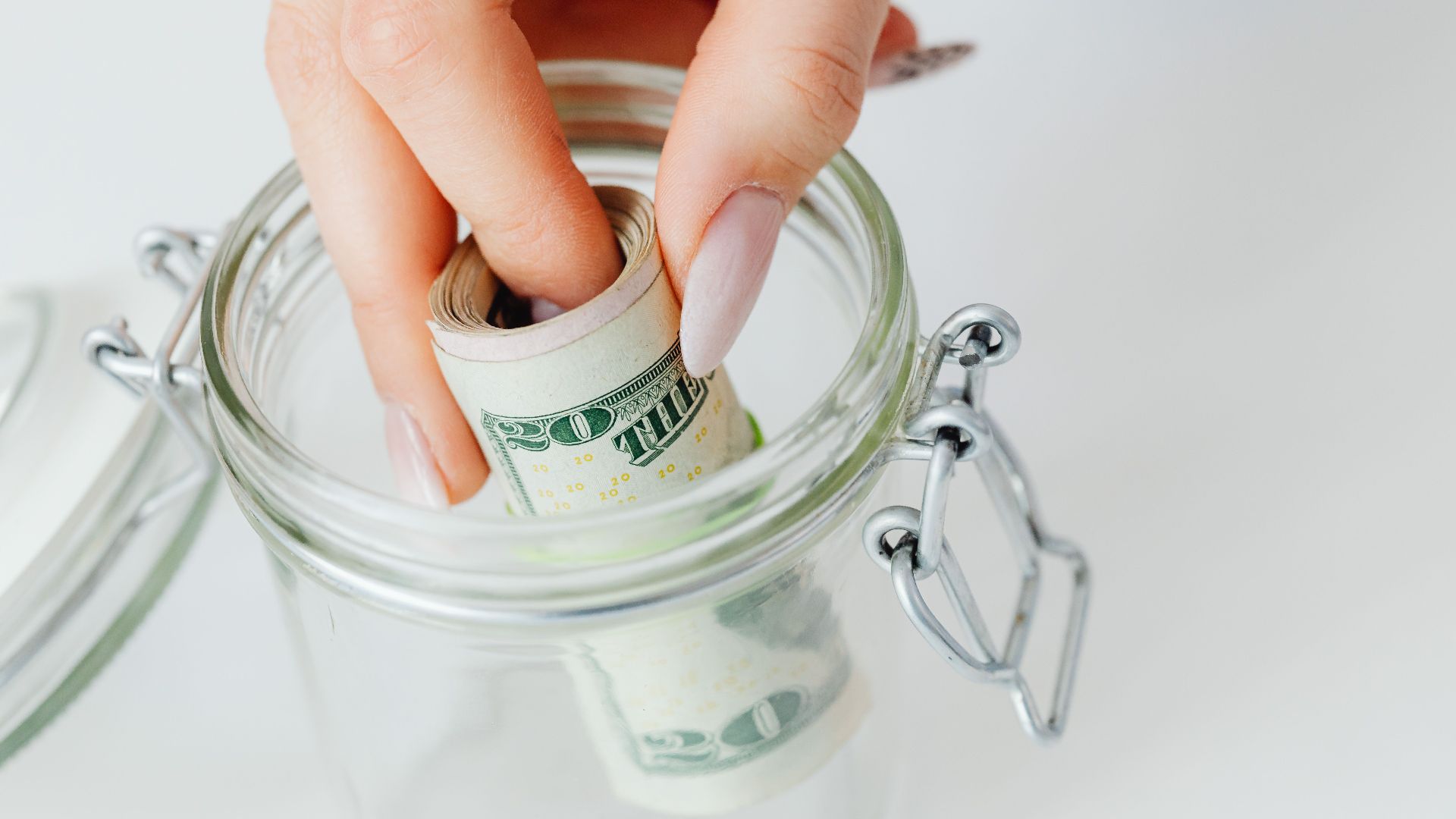 A close-up of a hand placing rolled dollars into a glass jar, symbolizing savings.