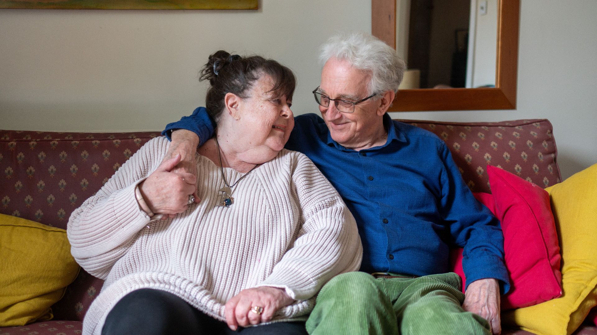 A loving senior couple smiling and embracing on a cozy sofa indoors.