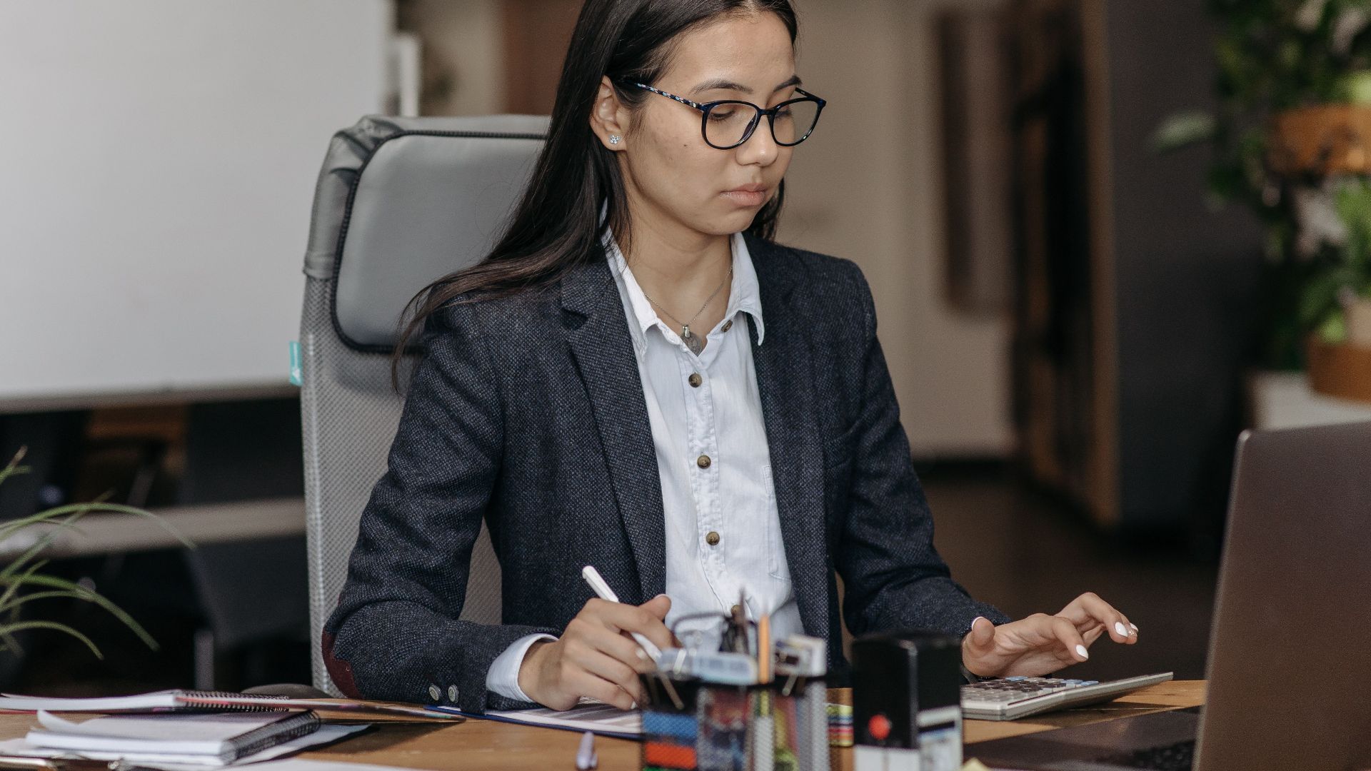 Young woman working with concentration at her laptop in a modern office setting, wearing eyeglasses.