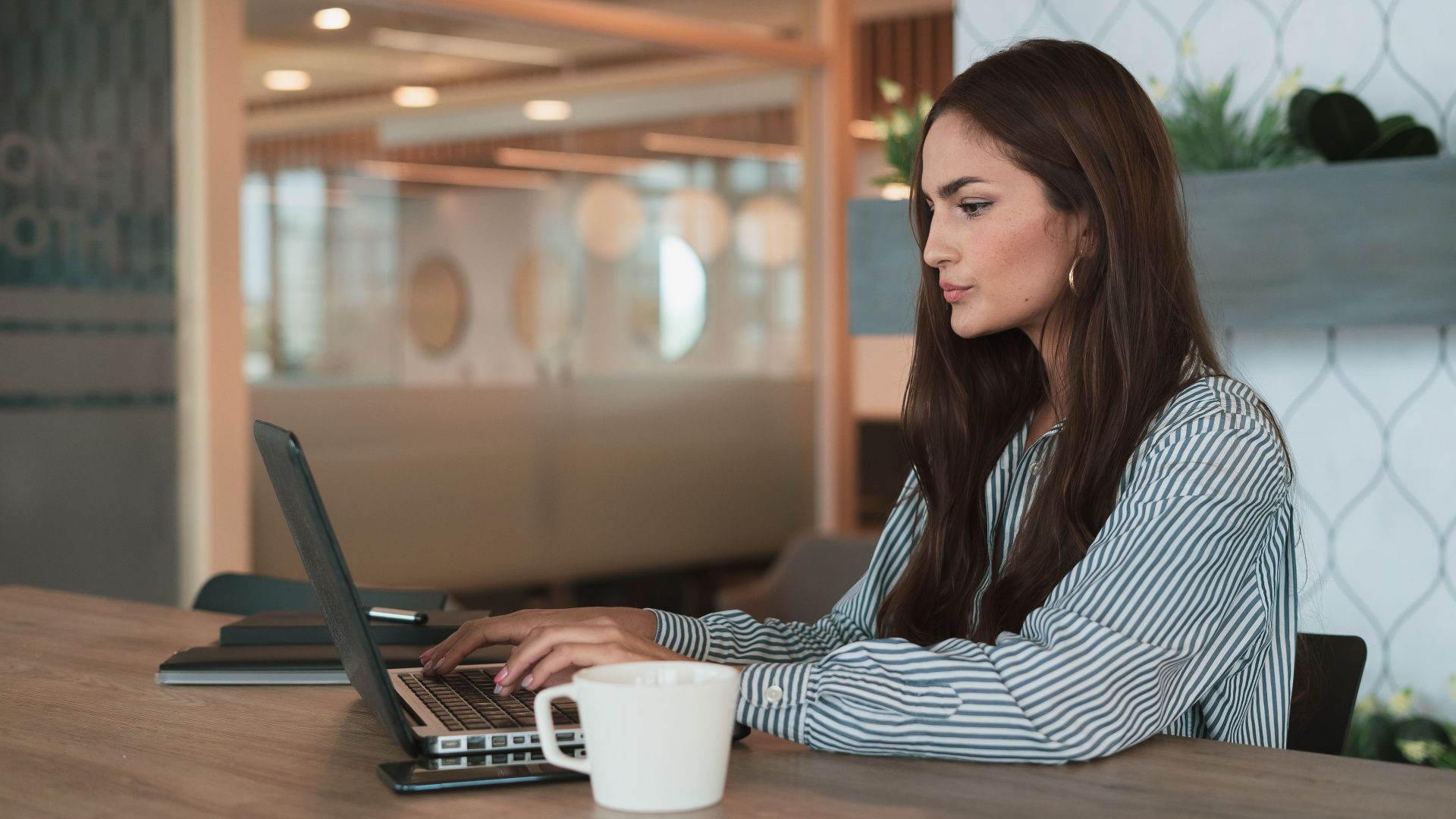 A professional woman working on her laptop at a modern office with a coffee cup nearby.