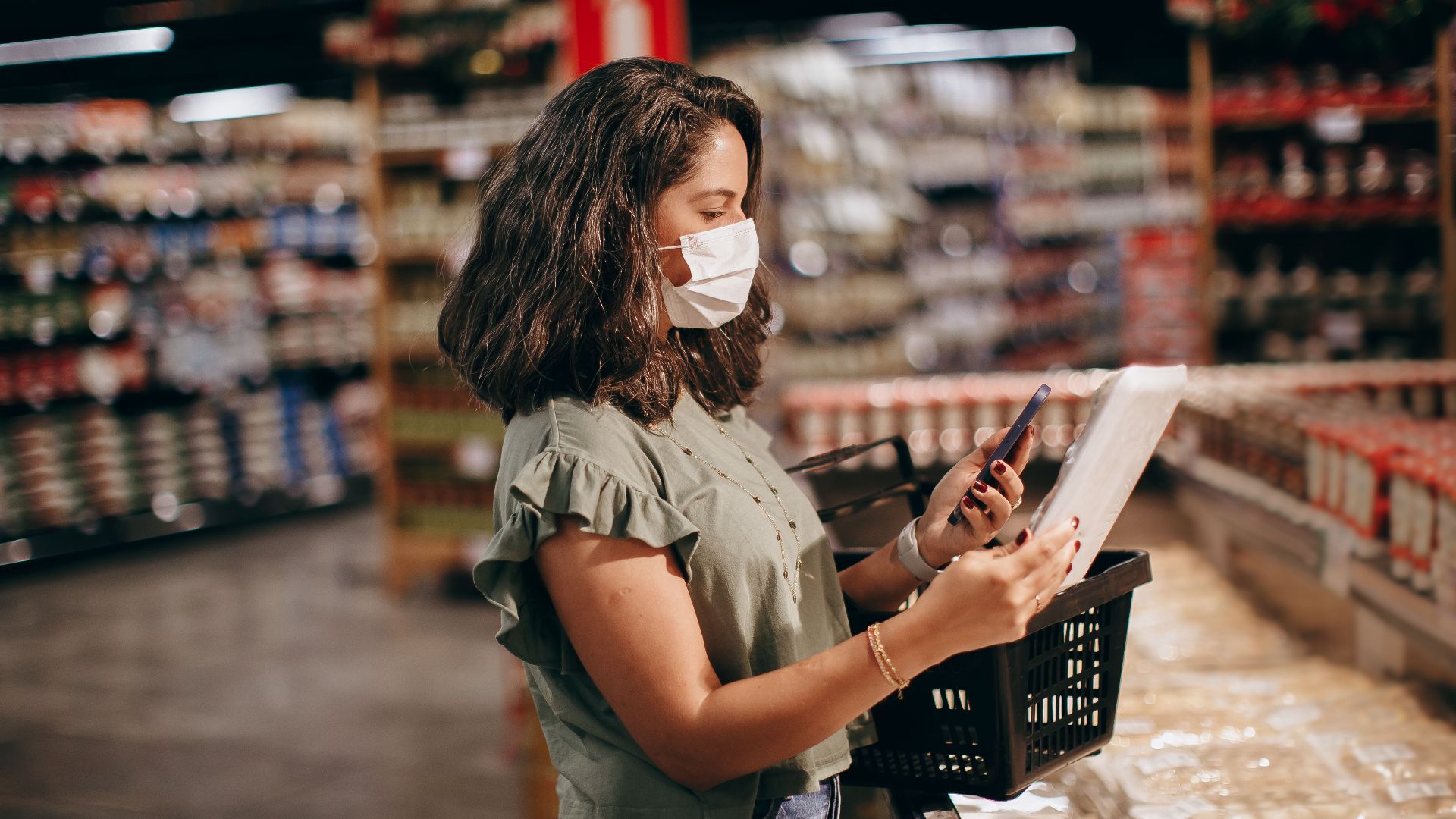 A woman shopping in a supermarket, wearing a face mask and using her phone to check products.