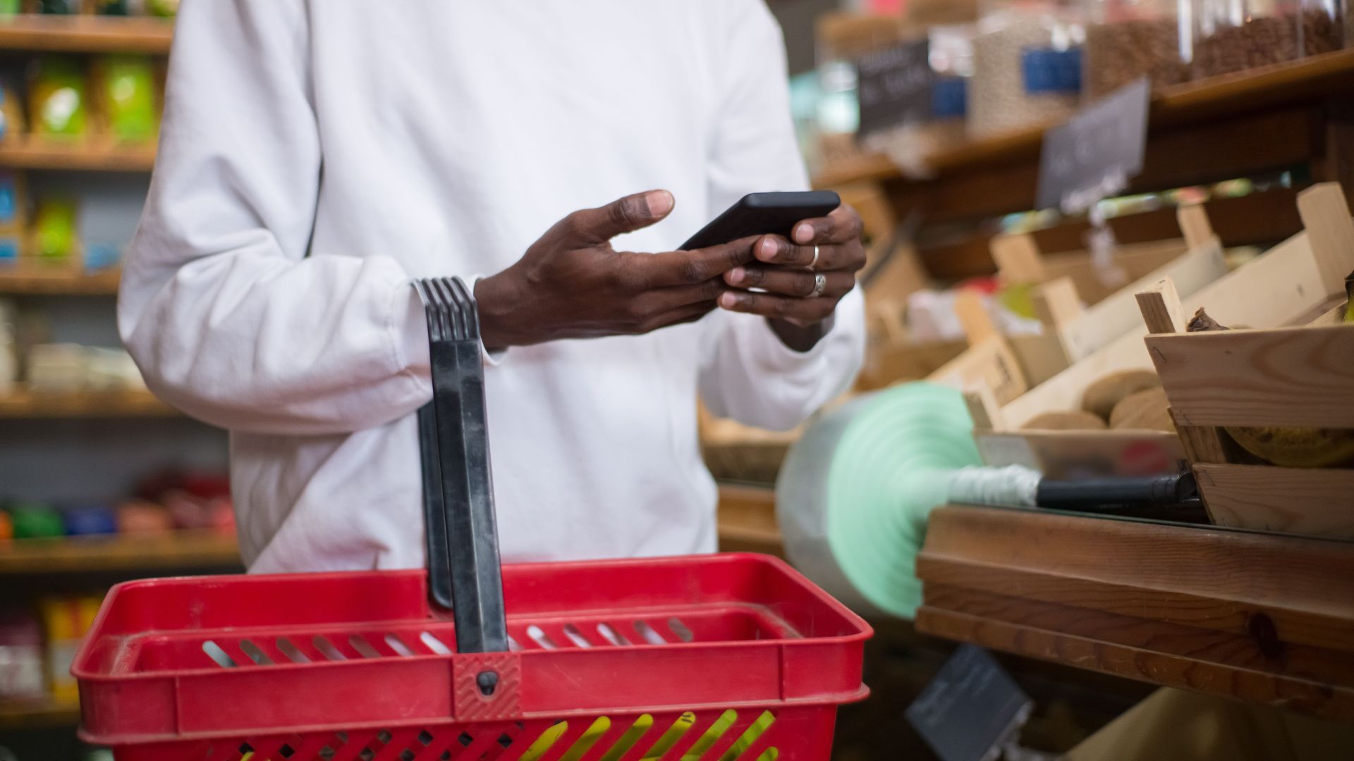 A man shops using a smartphone at a grocery store, holding a red basket filled with groceries.