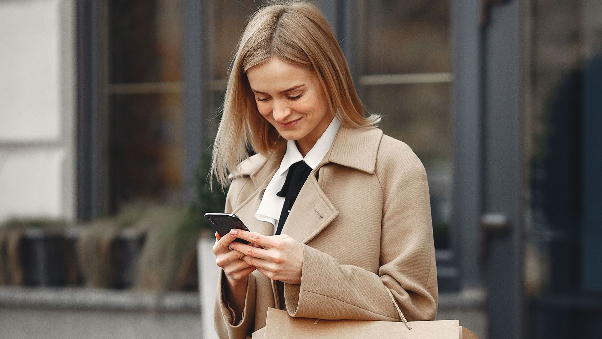 Positive elegant female in trendy coat carrying paper bags while using smartphone on street near restaurant entrance