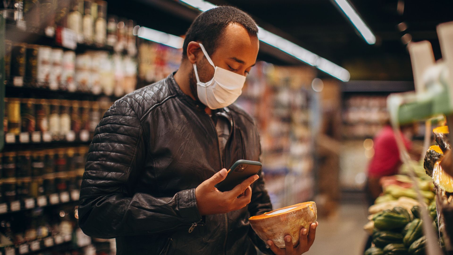 Man in leather jacket shopping with smartphone and mask in a grocery store aisle.