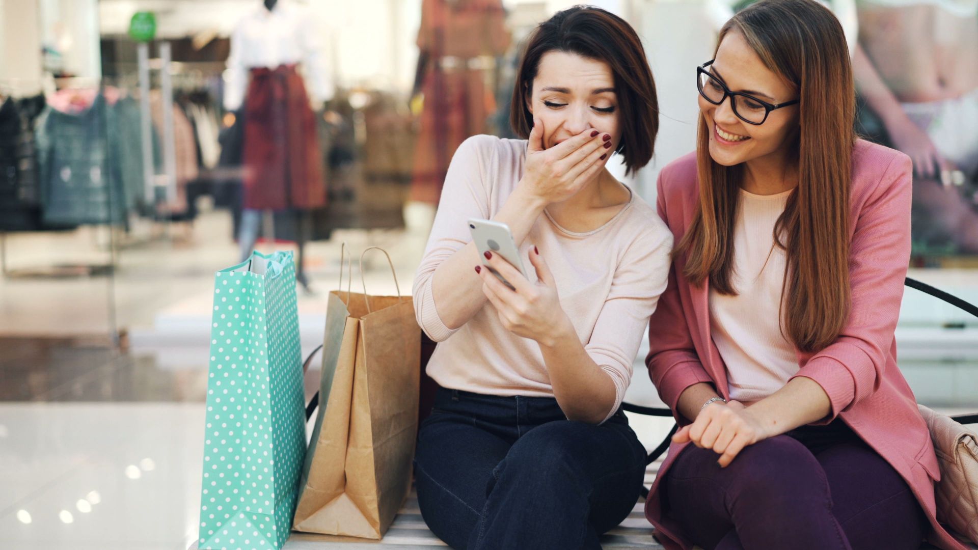 Two women sitting, laughing and looking at a phone in a shopping mall, surrounded by bags.