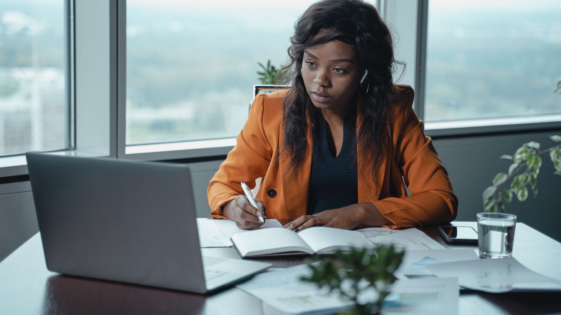 A businesswoman in a modern office intently working on a laptop and writing in a notebook.