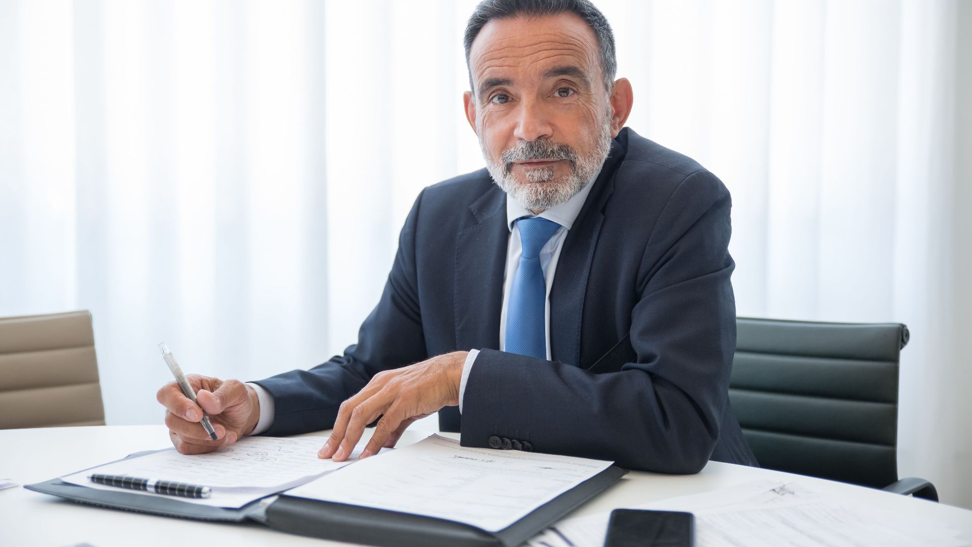 Elderly businessman with a beard signing documents in a modern office setting.