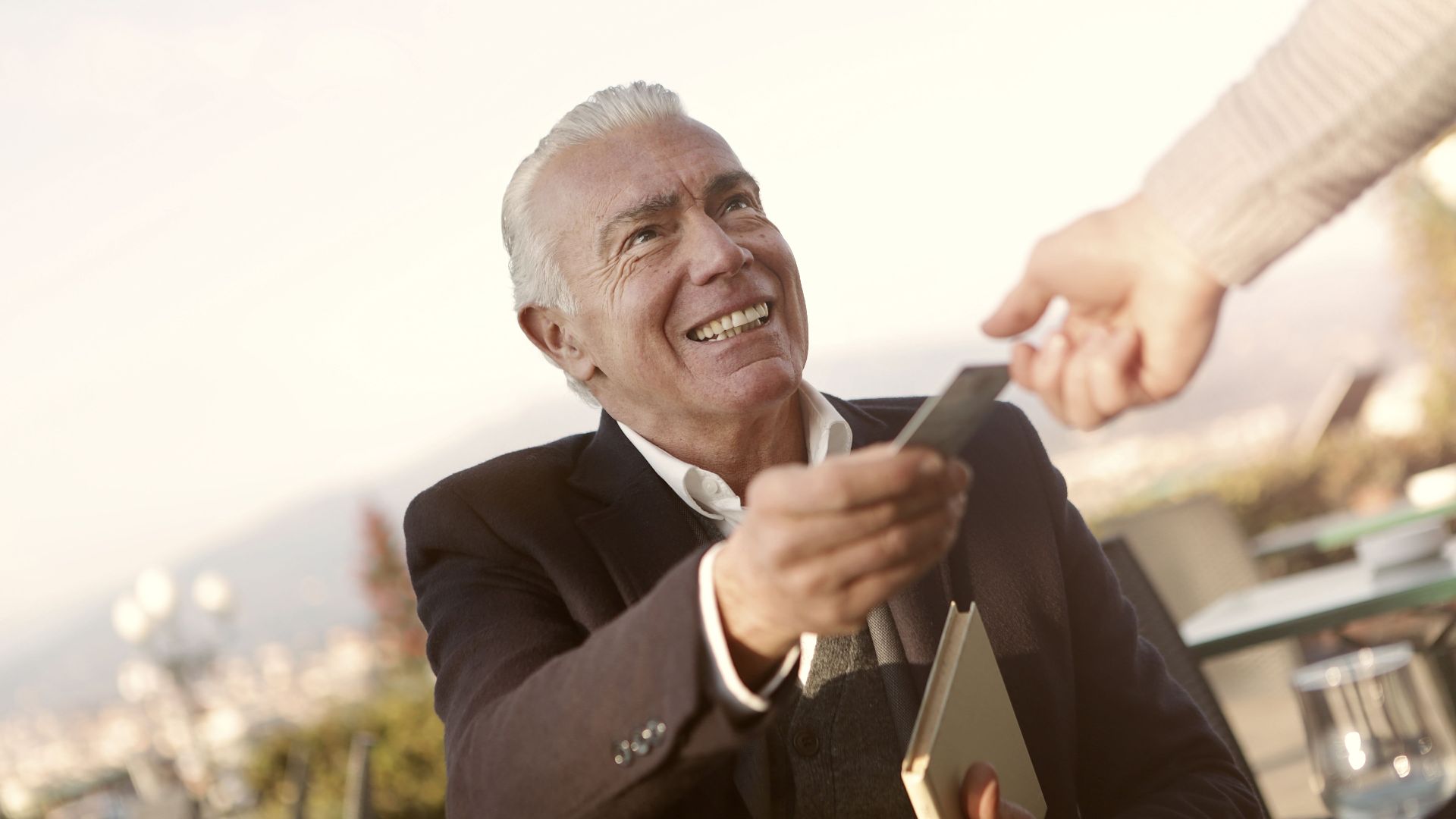 A senior man smiles as he hands a credit card outdoors, embodying trust and interaction.